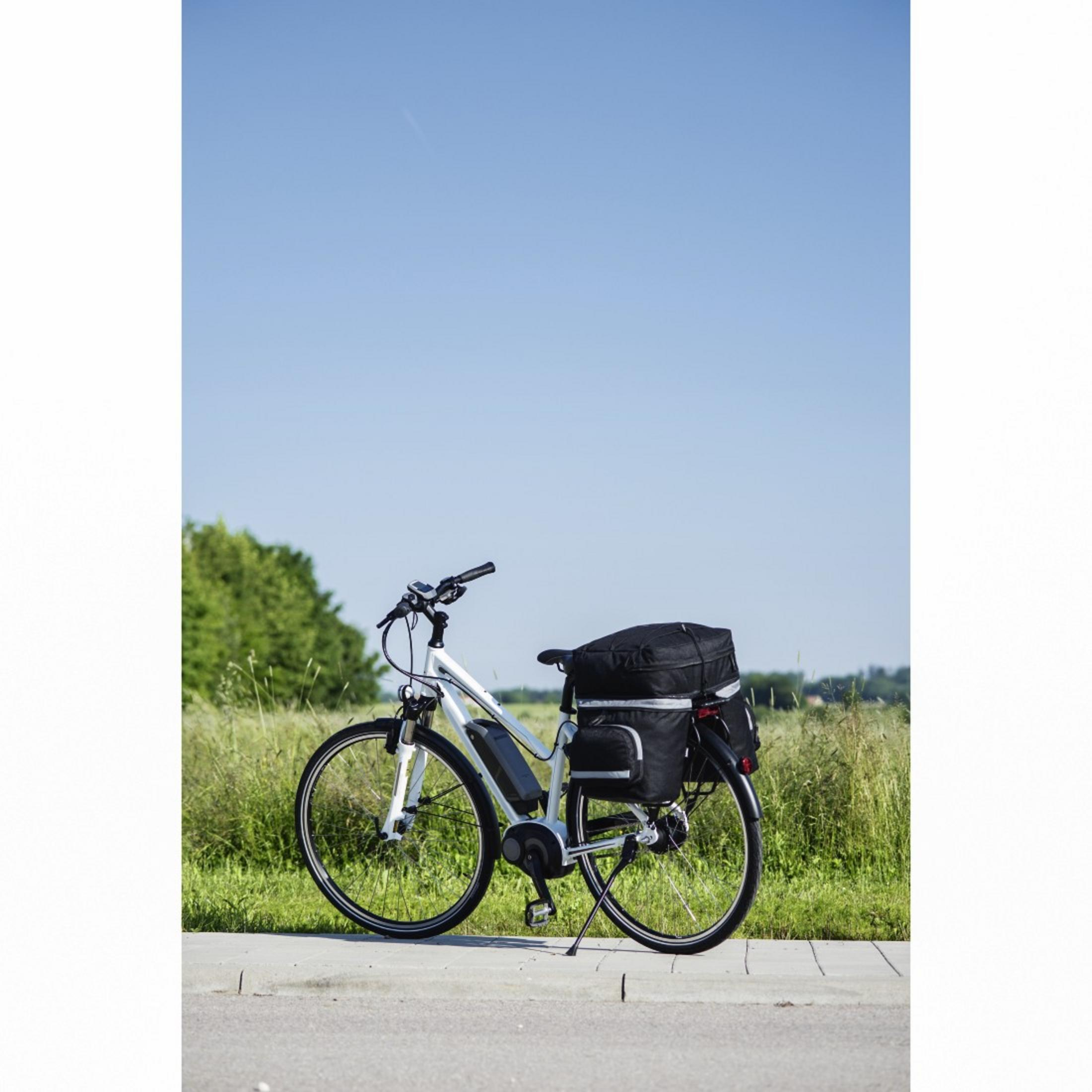 Ein weißes E-Bike mit schwarzen Gepäcktaschen steht auf einem Gehweg vor einer grünen Wiese unter klarem blaume Himmel.