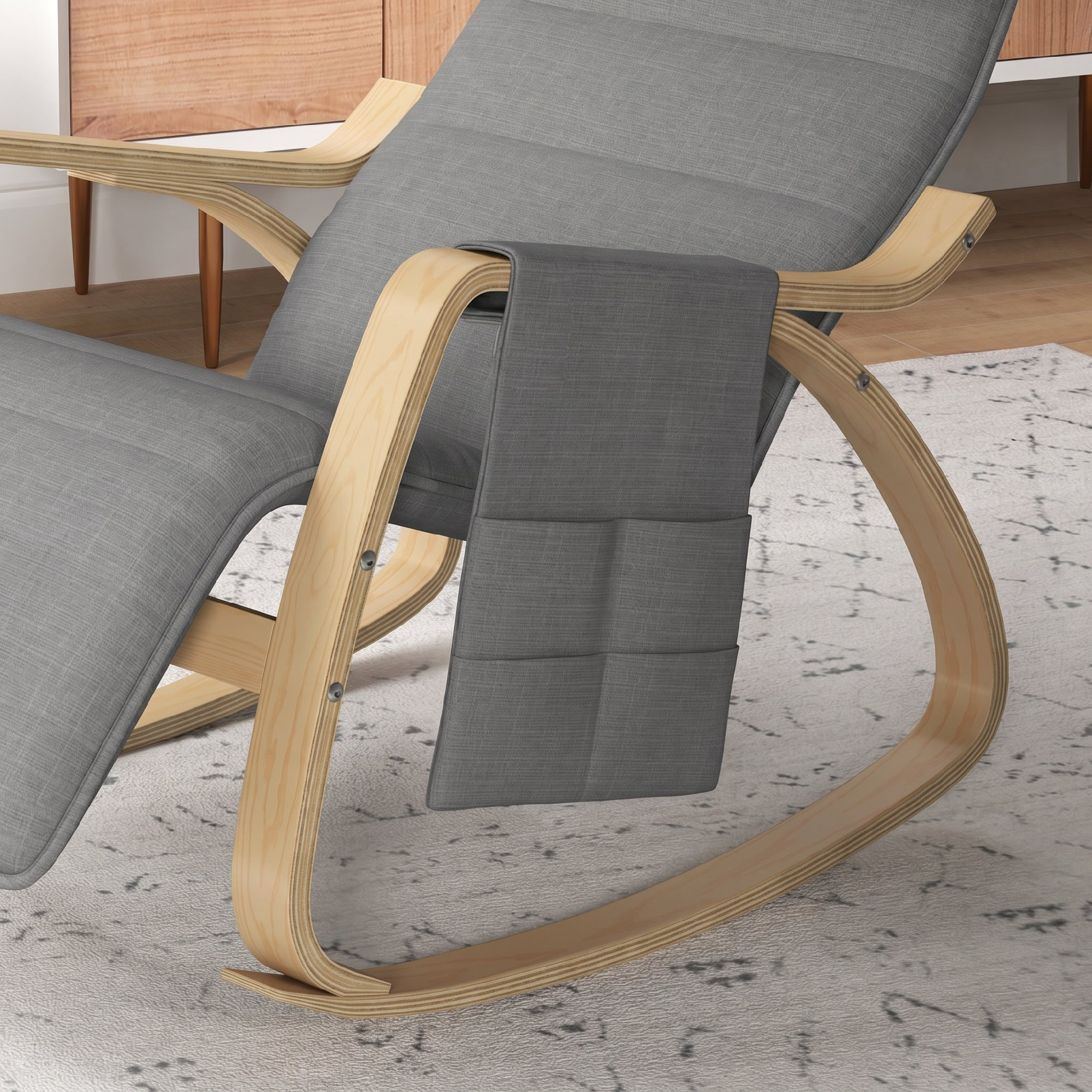 Mecedora gris con estructura de madera y bolsillo lateral sobre alfombra clara frente a un mueble de madera.