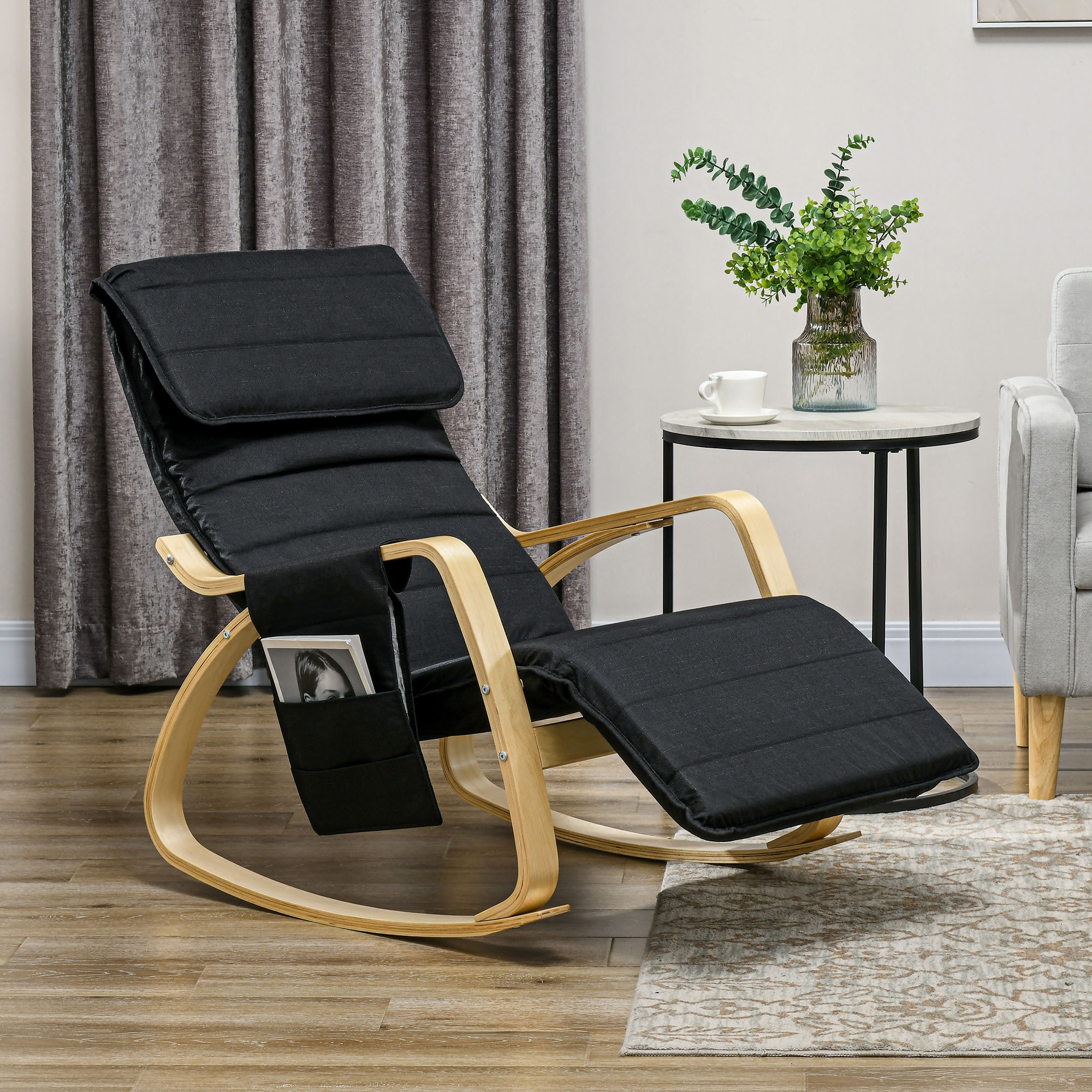 Una mecedora negra con estructura de madera y bolsillo lateral en una habitación con cortinas y mesa auxiliar.