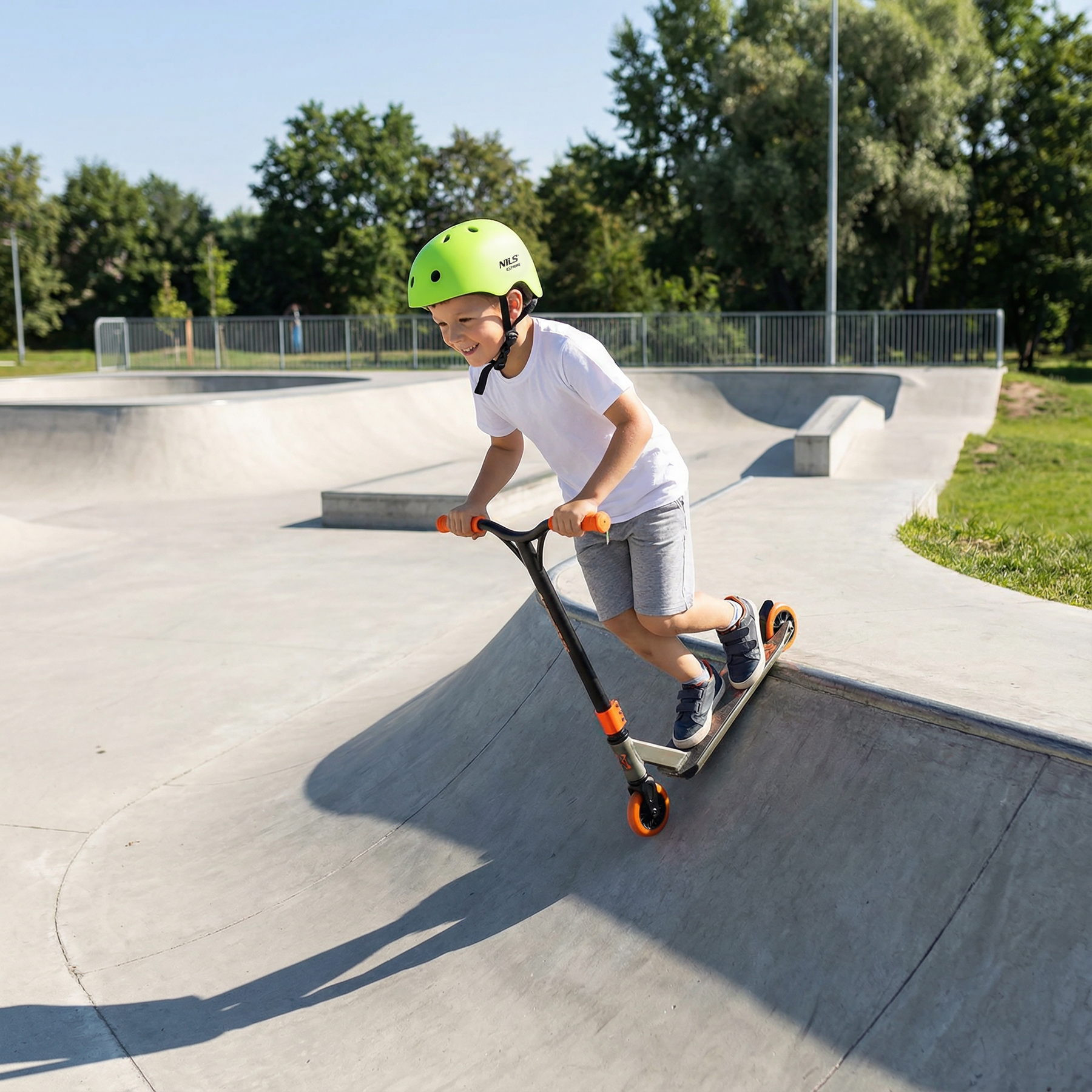 Młody chłopiec w zielonym kasku jezdzi na hulajnodze po betonowej rampie w skateparku w słoneczny dzien.