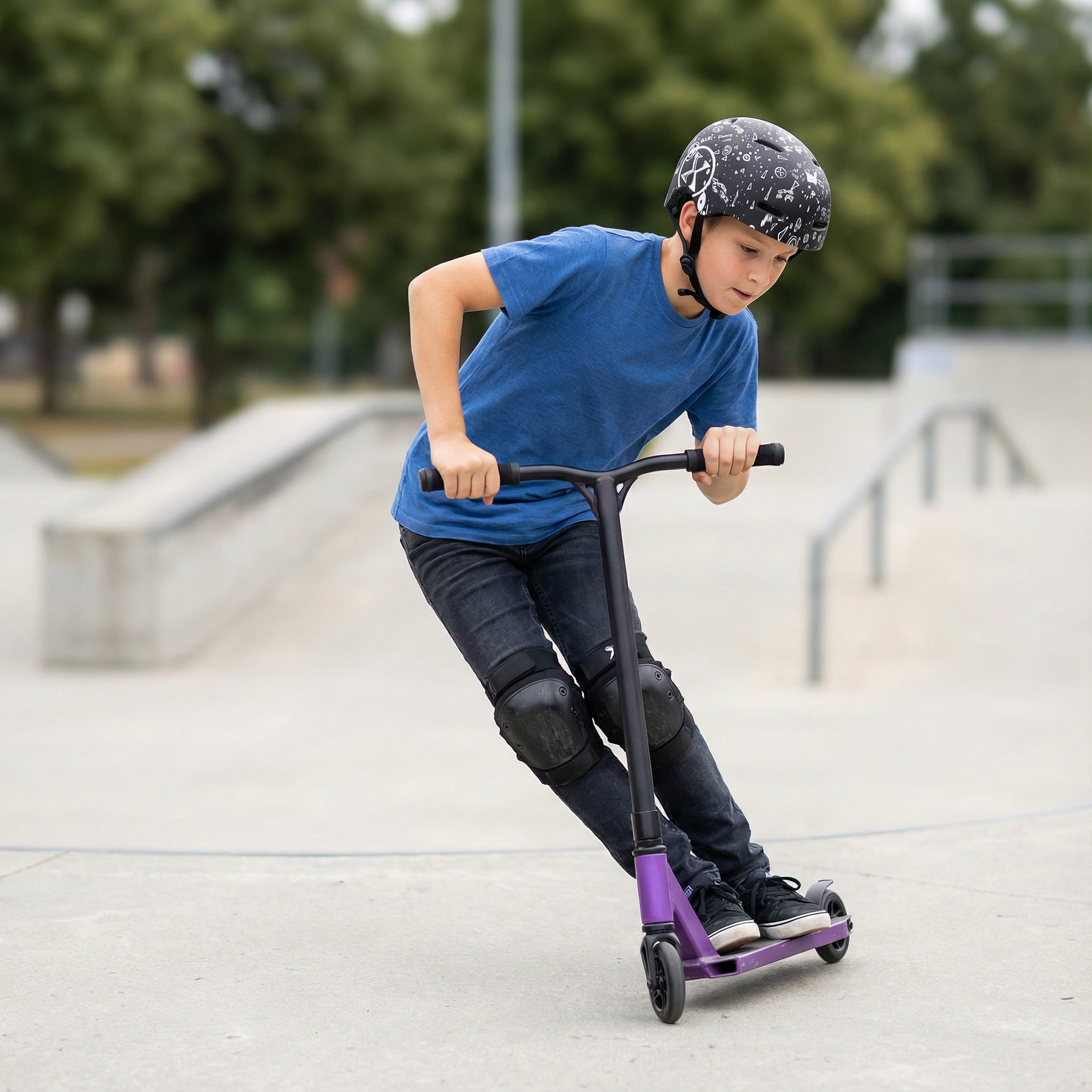Chłopiec w niebieskiej koszulce i kasku jeździ na hulajnodze wyczynowej w skateparku.