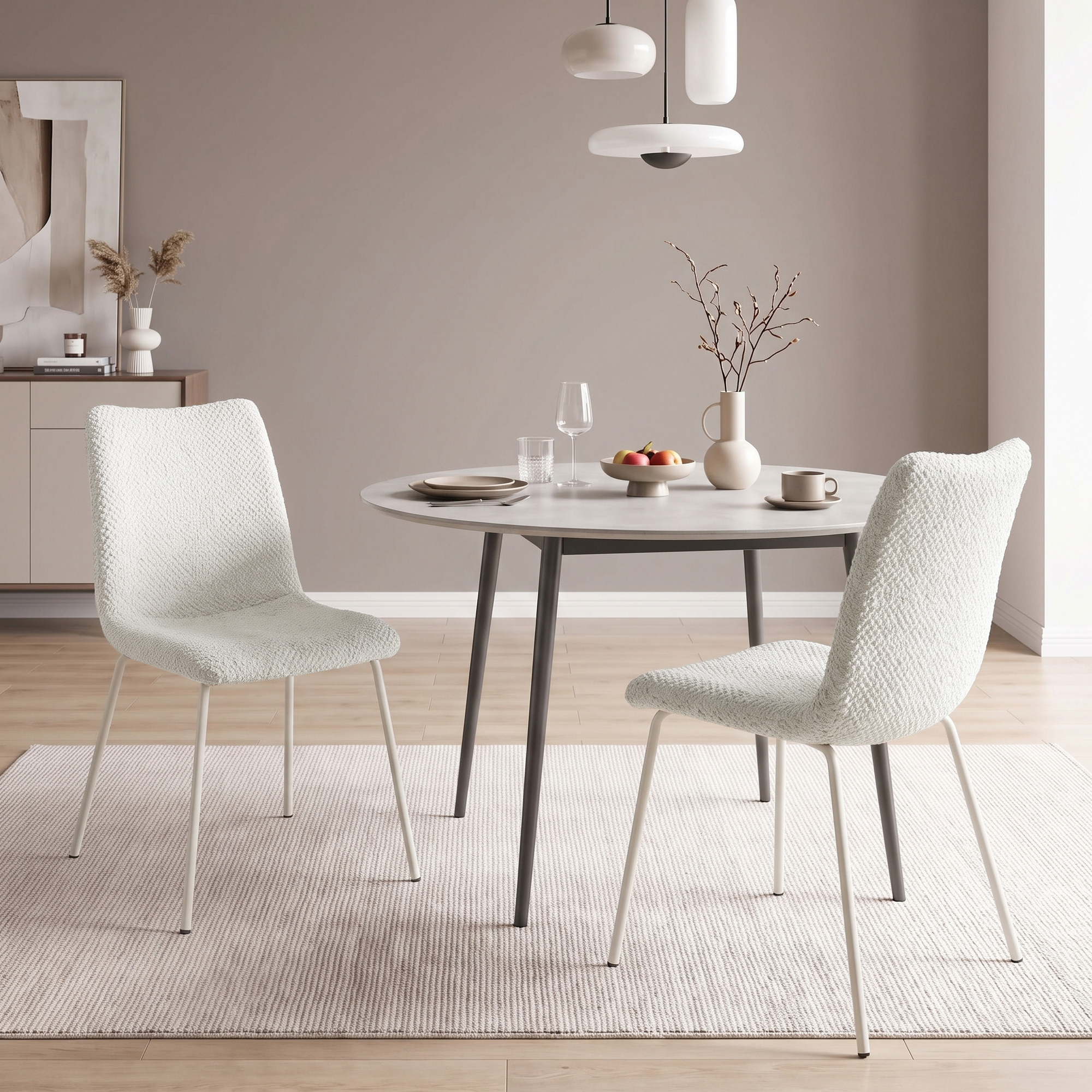 Comedor moderno con dos sillas blancas, mesa redonda, vajilla, florero y lámpara colgante en una habitación neutral.