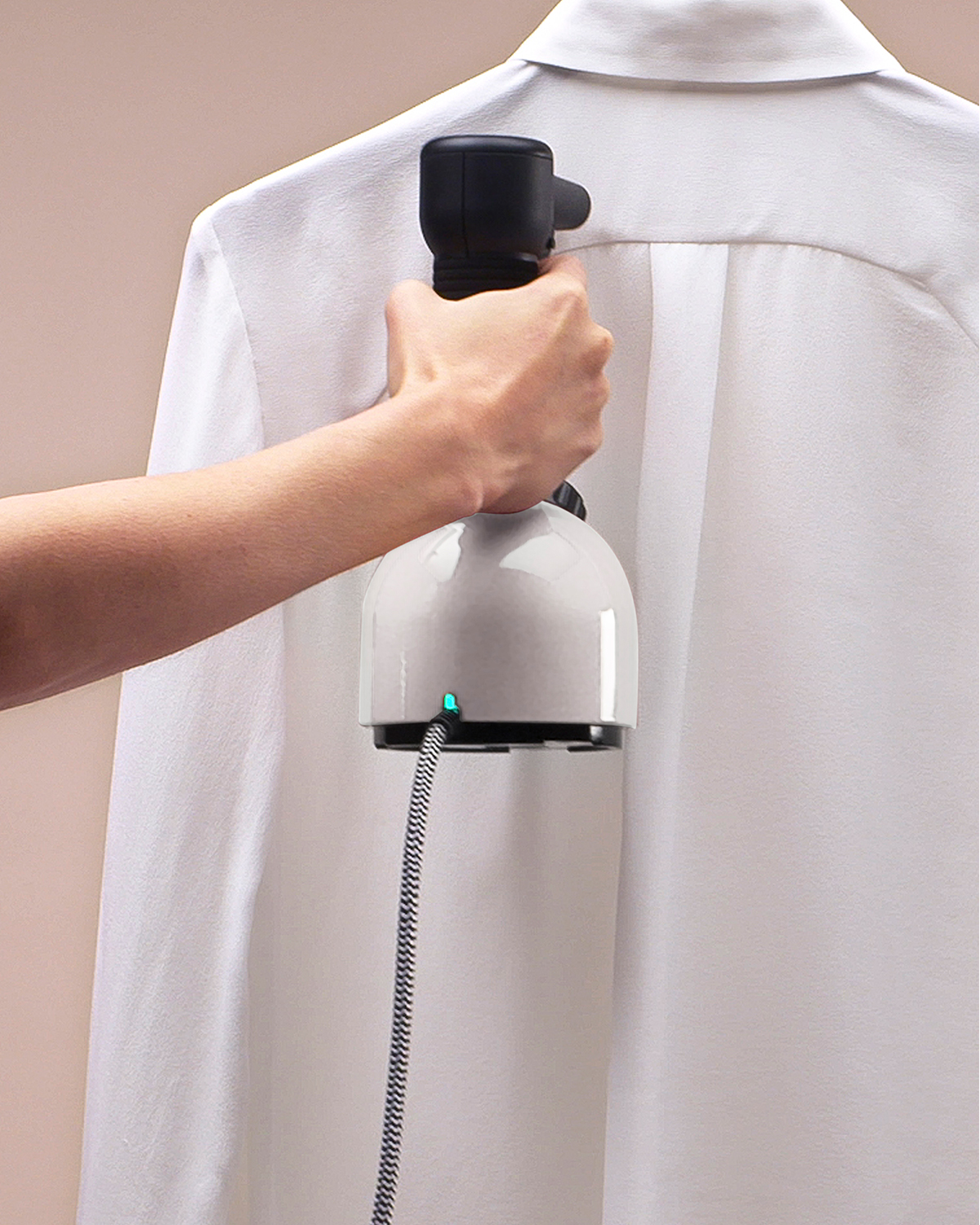 Una mano tiene un vaporizzatore portatile bianco e nero, stirando la schiena di una camicia bianca appesa.
