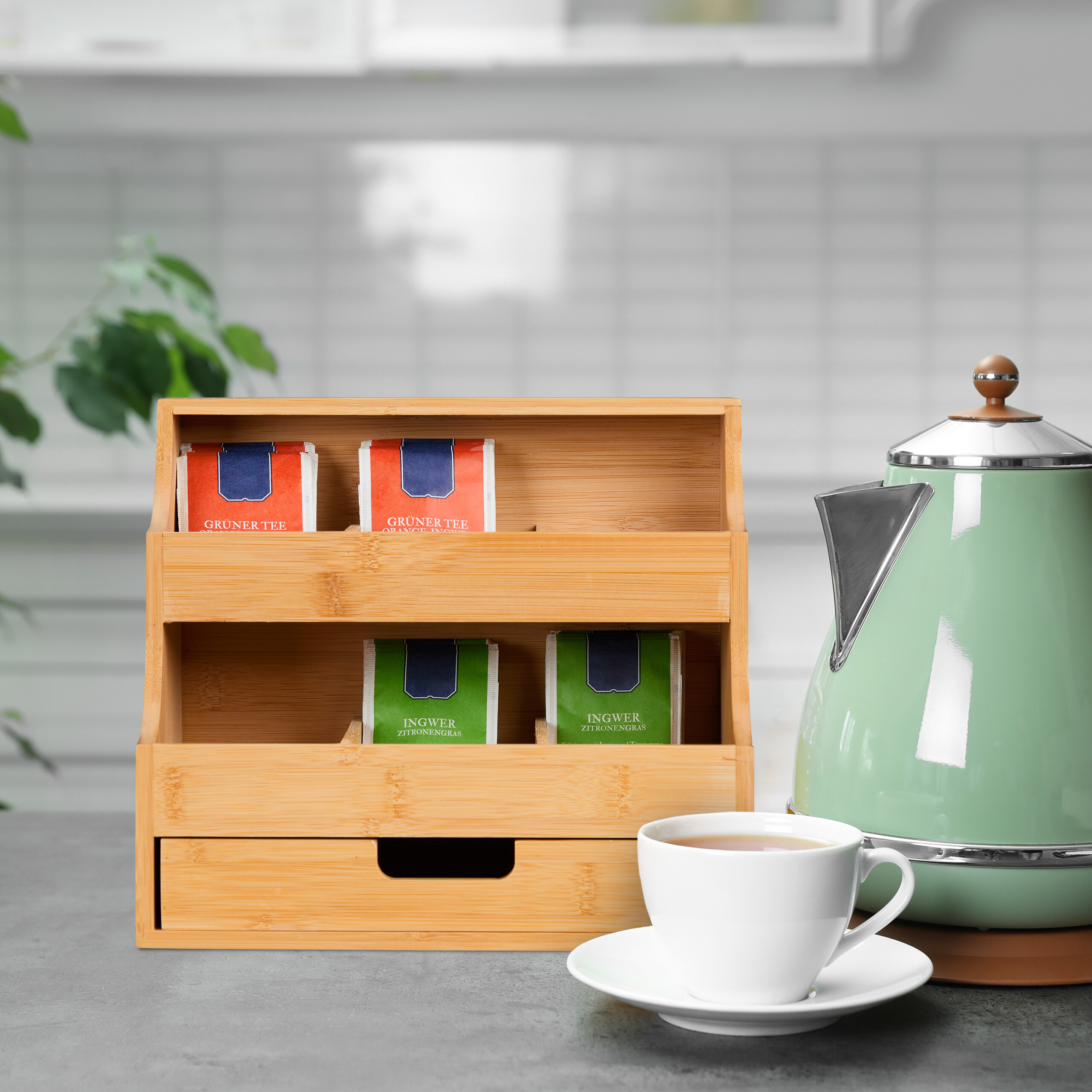 Bambus-Tee-Organizer, grüne Teekanne und eine Tasse Tee auf einer grauen Arbeitsplatte in einer Küche.