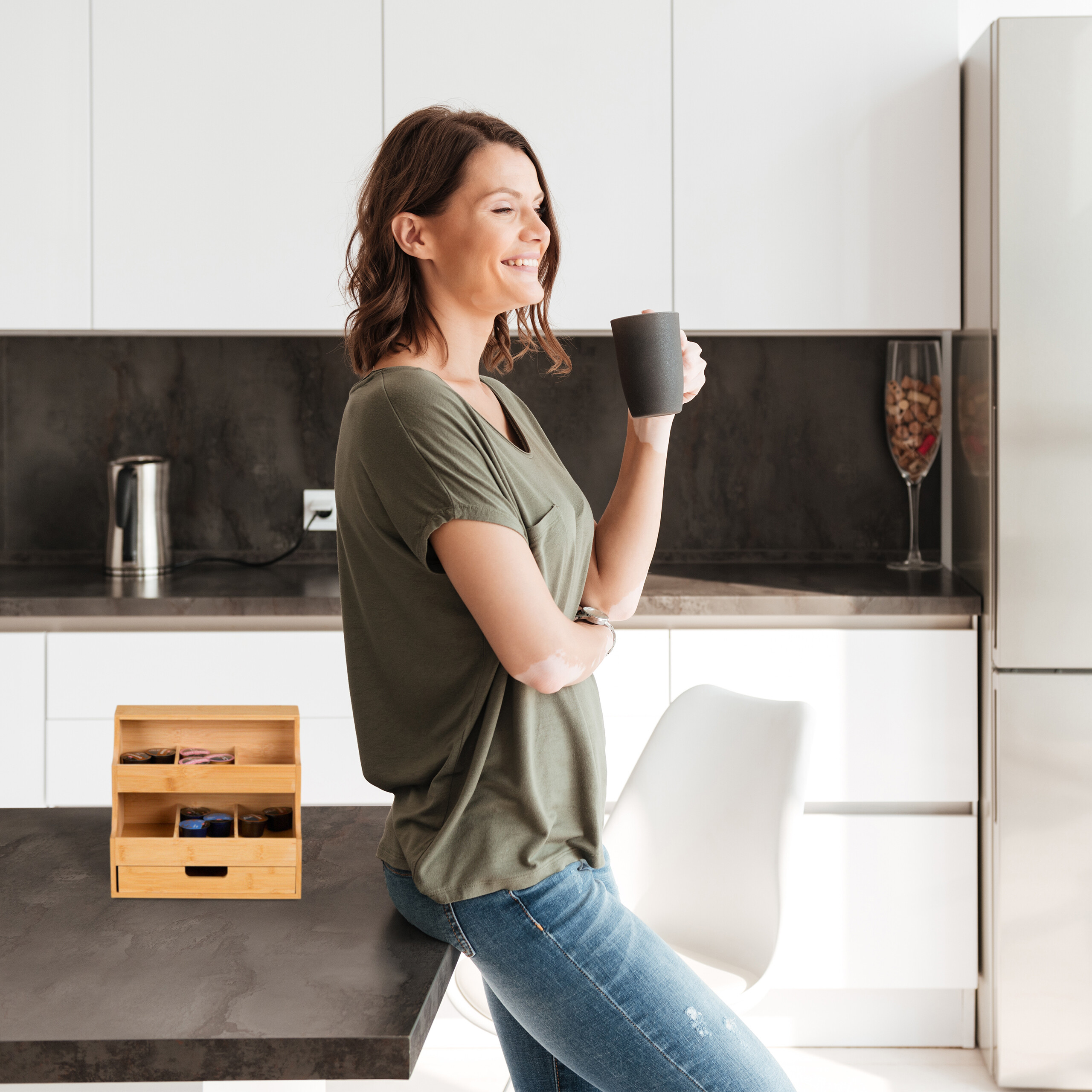 Eine lächelnde Frau hält eine dunkle Tasse in einer modernen weißen Küche mit einem Kaffeekapselhalter auf dem Tresen.