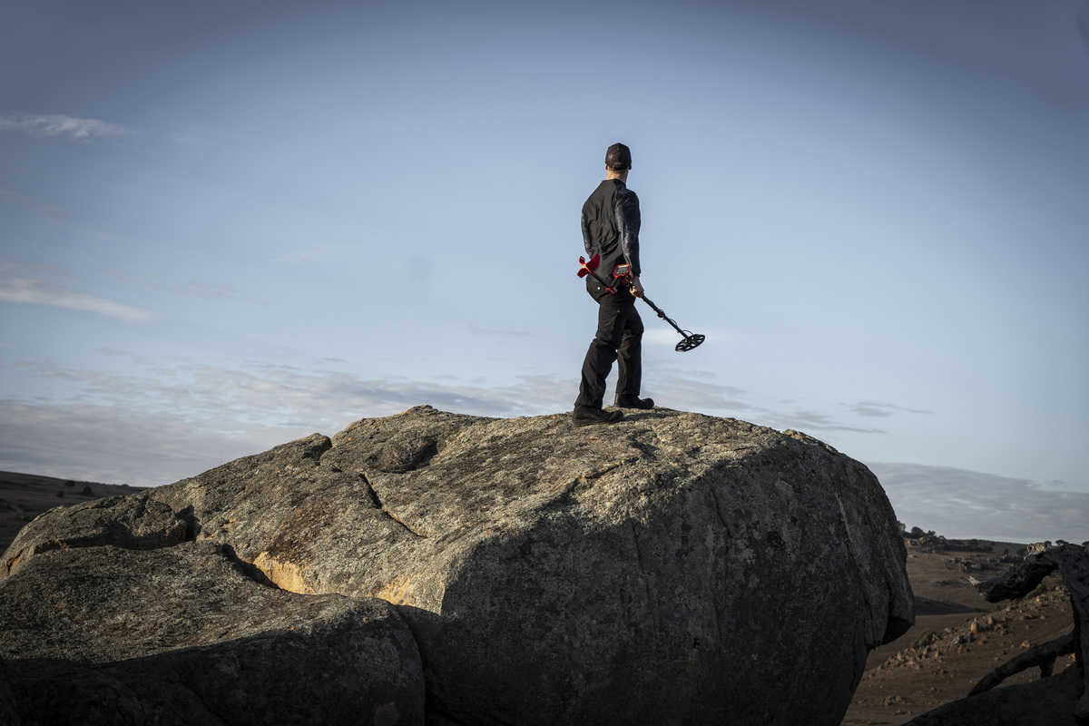 Un hombre con un detector de metales está en la cima de una gran roca bajo un cielo azul.