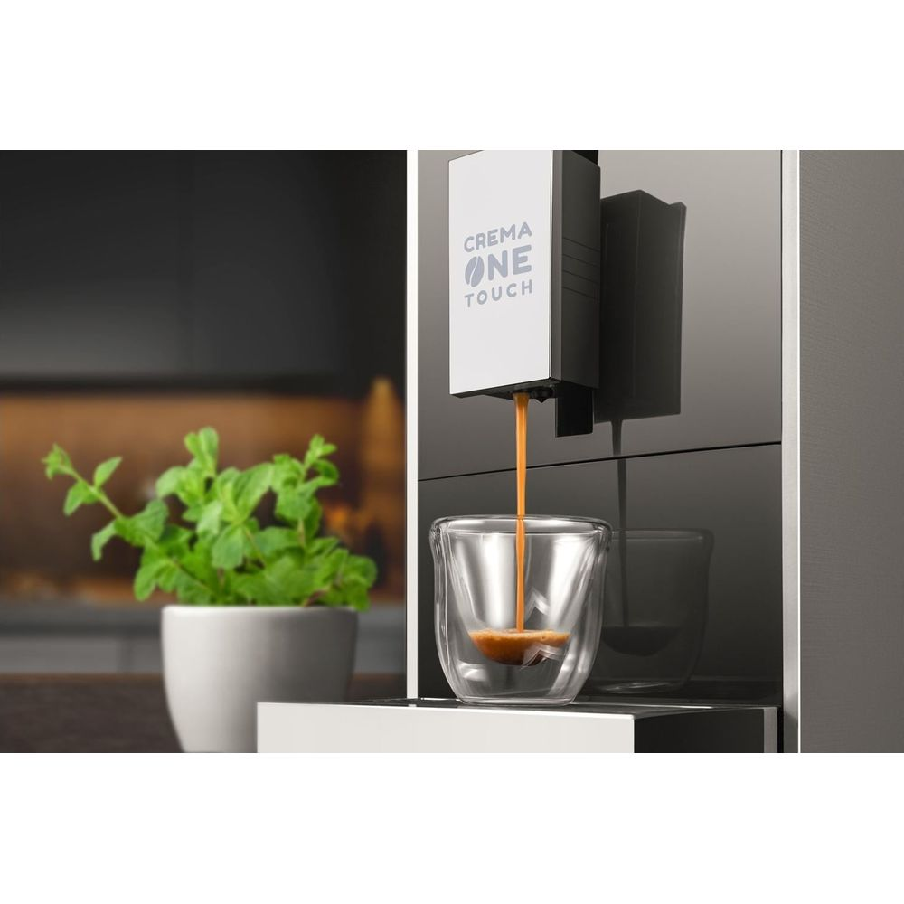 Una macchina da caffè etichettata CREMA ONE TOUCH versa espresso in un bicchiere piccolo. Sullo sfondo c'è una pianta.