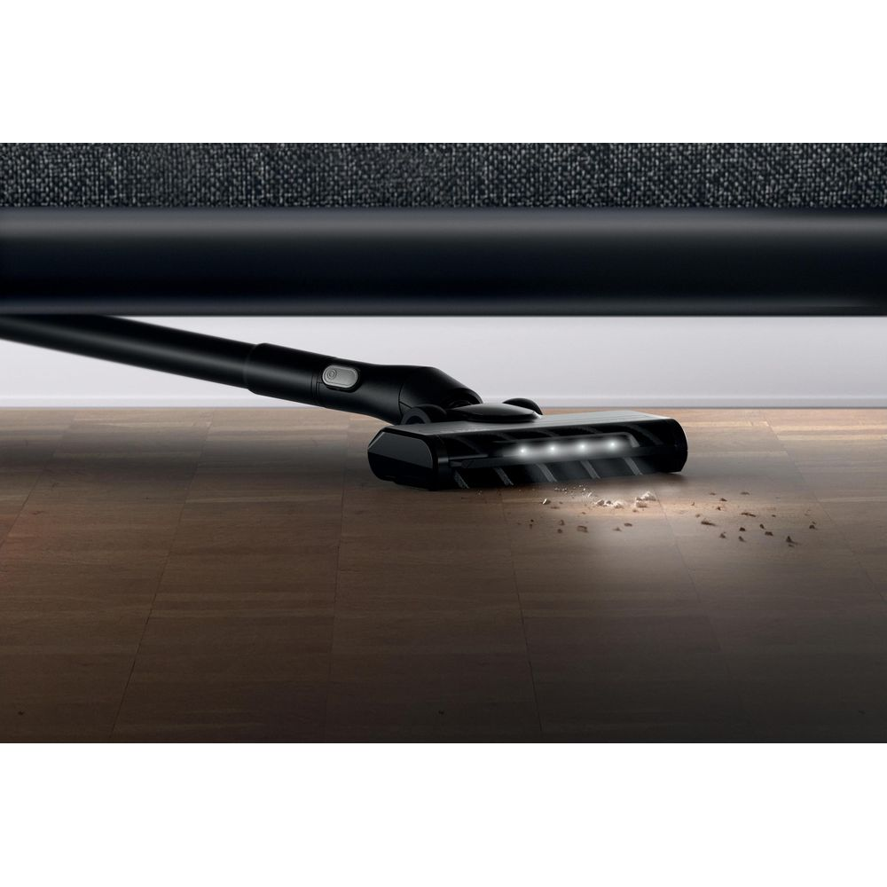 Un aspirapolvere nero con luci LED pulisce i detriti su un pavimento in parquet sotto un mobile.