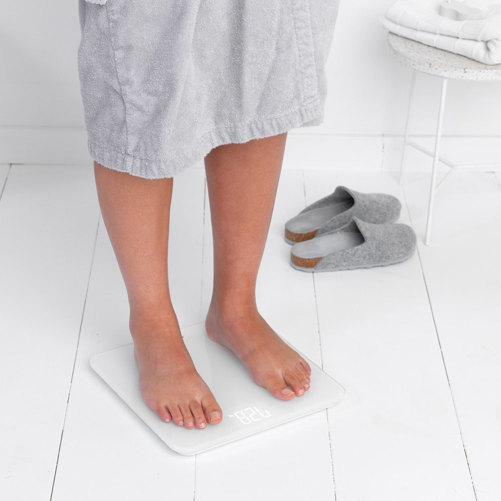 Persona con accappatoio grigio in piedi su una bilancia bianca su pavimento in legno bianco.