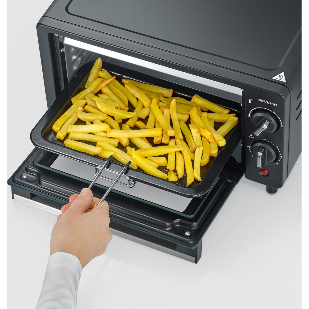Eine Hand nimmt mit einer Zange ein Backblech mit Pommes frites aus einem kleinen schwarzen Ofen.