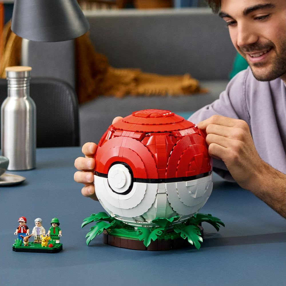 Un hombre construye una gran Pokéball de Lego en una mesa azul, con pequeñas figuras al lado.