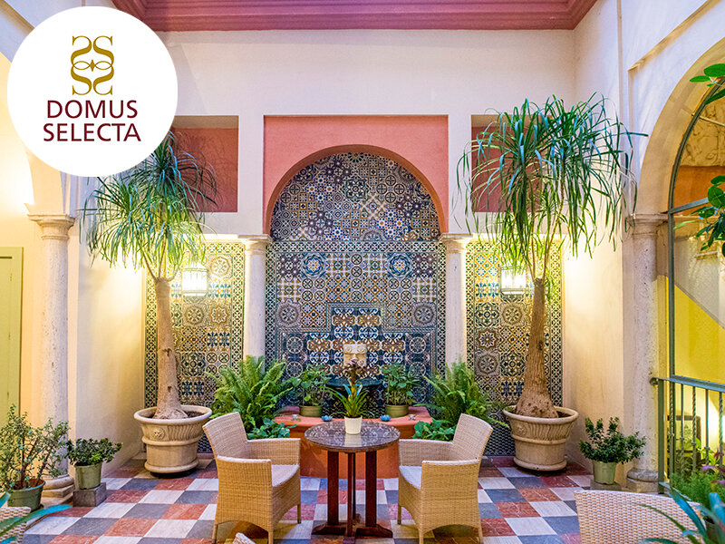 Un patio interior con azulejos estampados, columnas, dos sillones de mimbre, una mesa y grandes macetas.