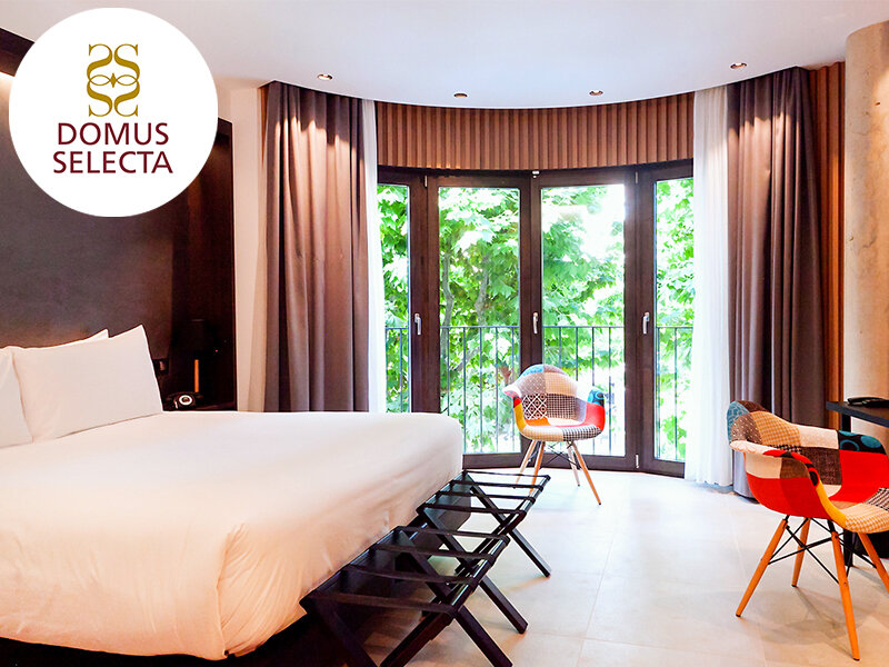 Habitación de hotel moderna con cama grande, sillones coloridos y ventanales con vista a la vegetación.