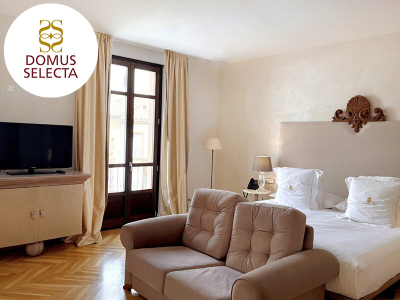 Una habitación de hotel con cama, sofá marrón, televisor y puerta al balcón con cortinas beiges.