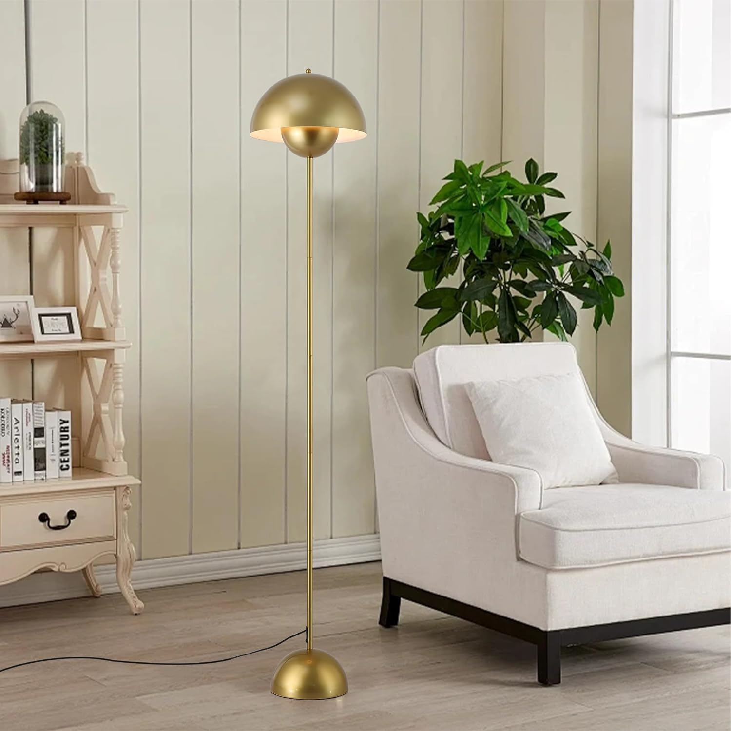 Una lámpara de pie dorada en una sala con sillón blanco, estante y planta verde frente a una pared.