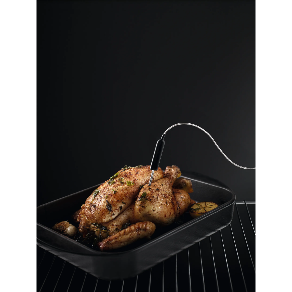 Un pollo asado en un plato negro con un termómetro para carne en una rejilla del horno.