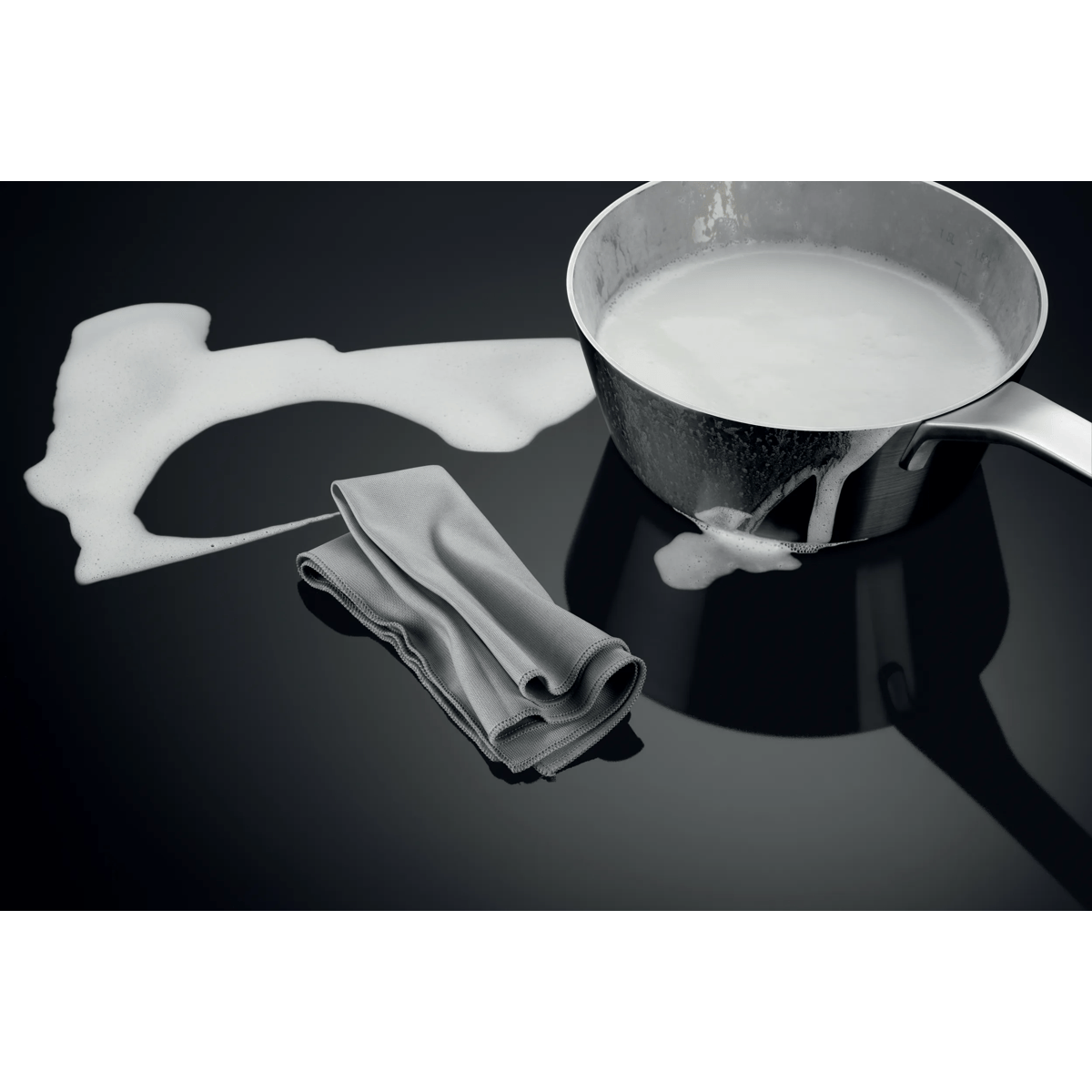 Una olla con leche en la placa junto a un derrame de espuma y un paño gris.