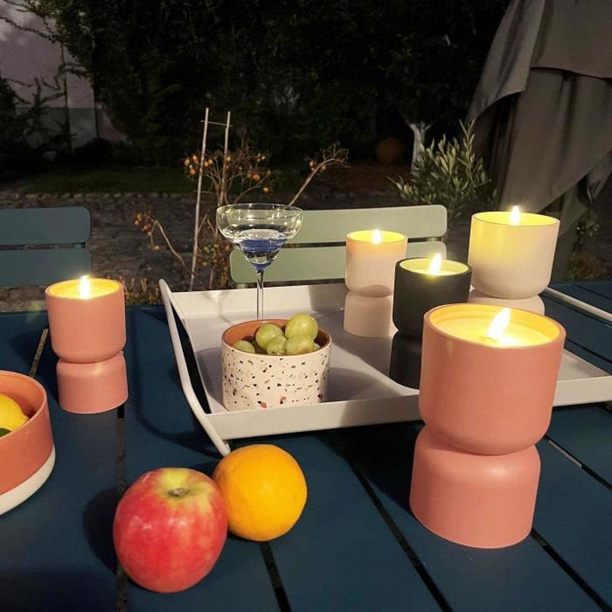 Beleuchtete Kerzen, ein Glas, Obst und ein Tablett stehen abends auf einem Gartentisch.