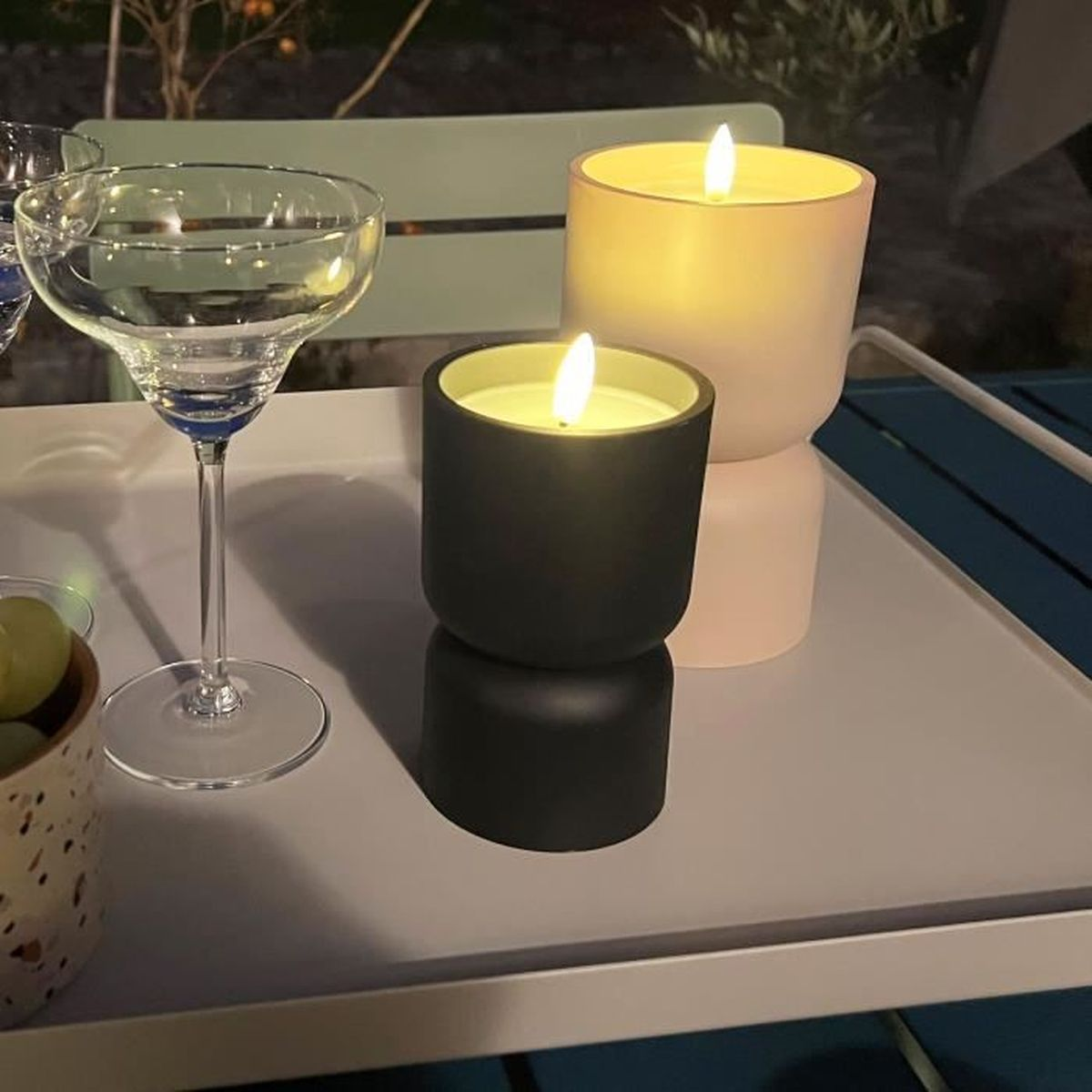 Zwei brennende Kerzen in modernen Haltern und ein leeres Glas stehen nachts draußen auf einem weißen Tablett.