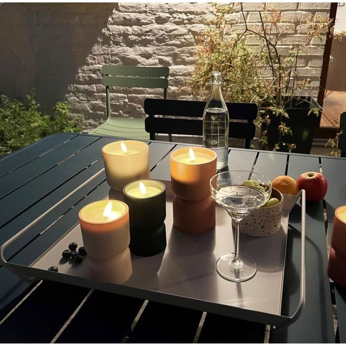 Ein Tablett mit Kerzen, einem Glas Wasser und Obst auf einem Tisch im Freien bei Abendlicht.