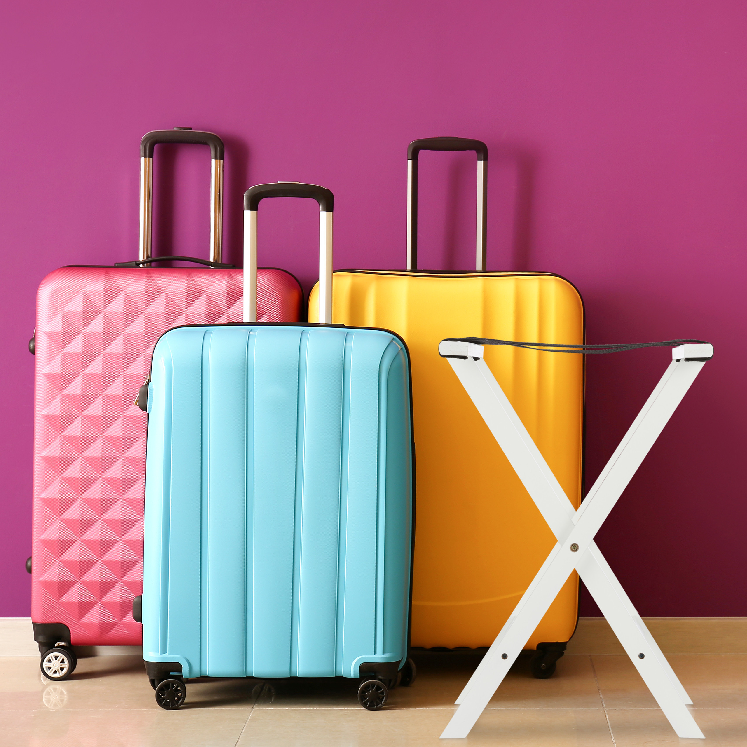 Drei bunte Koffer in Rosa, Blau und Gelb stehen vor einer lila Wand mit einem weißen Klappgepäckständer.