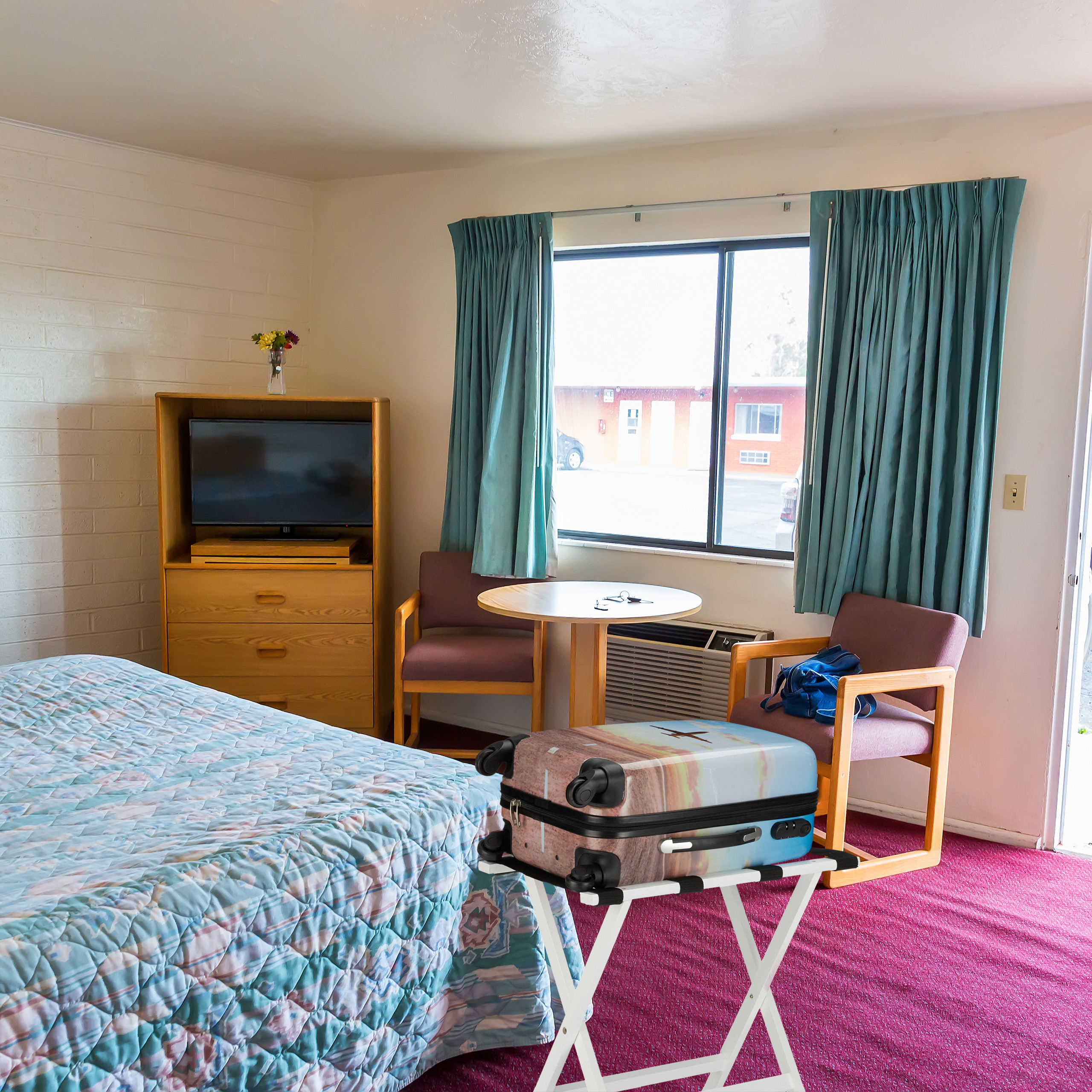 Ein Hotelzimmer mit Bett, Koffer auf Ständer, Fernseher, Fenster mit blauen Vorhängen und Sesseln.