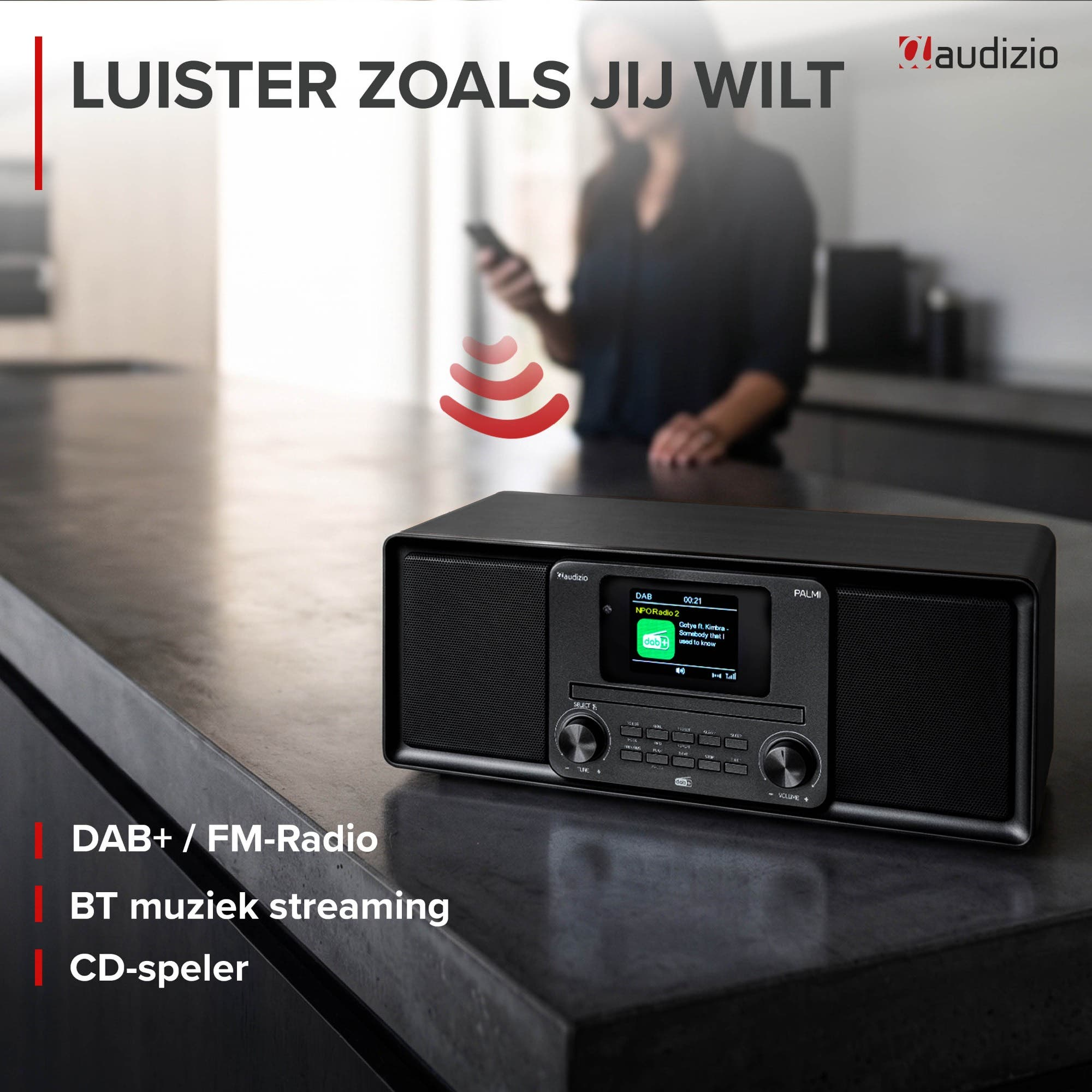 Ein schwarzes Audizio Radio auf einer Küchentheke mit einer unscharfen Frau im Hintergrund, die ein Smartphone bedient.