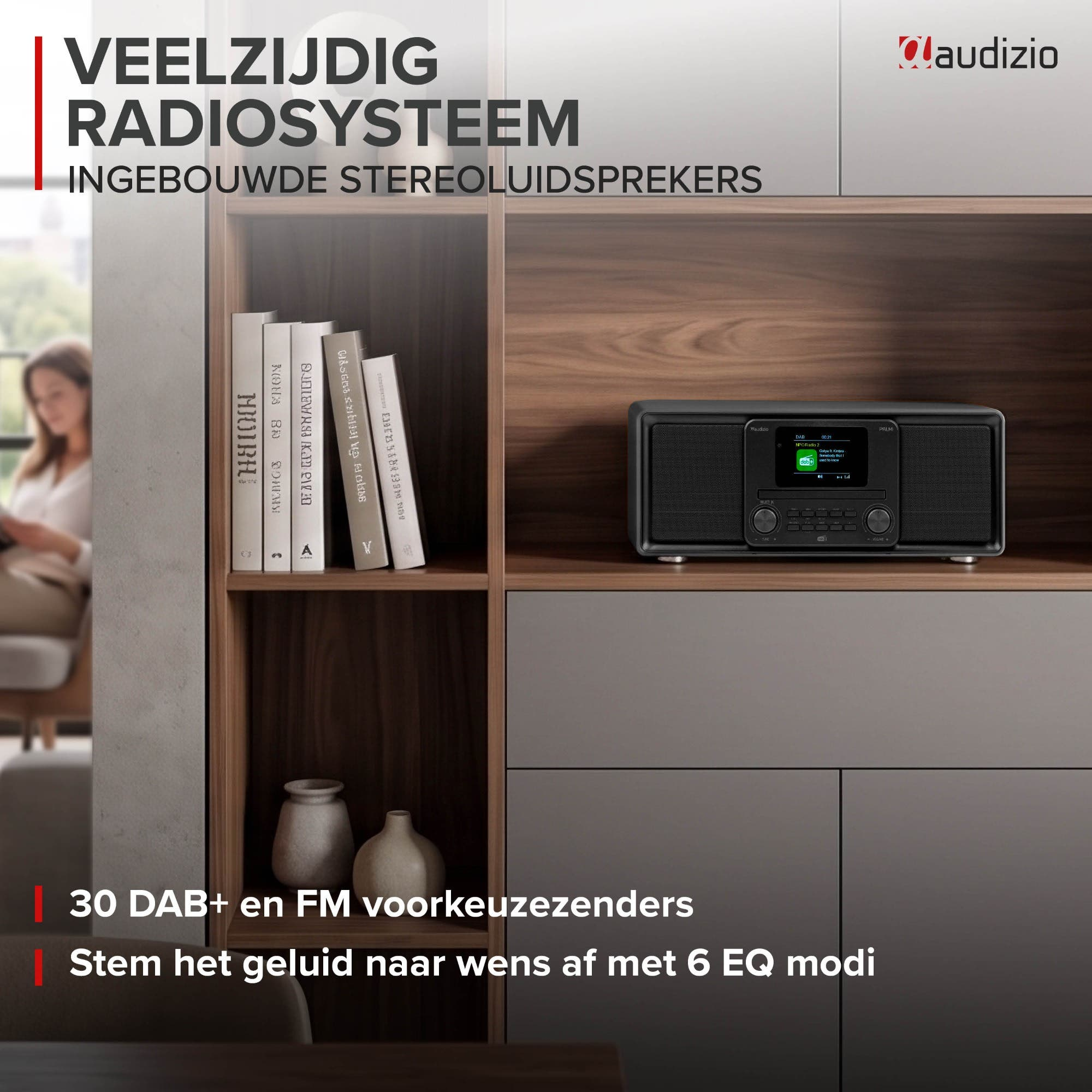 Ein schwarzes Audizio-Radio steht auf einem Holzregal neben Büchern und Vasen, im Hintergrund liest jemand.