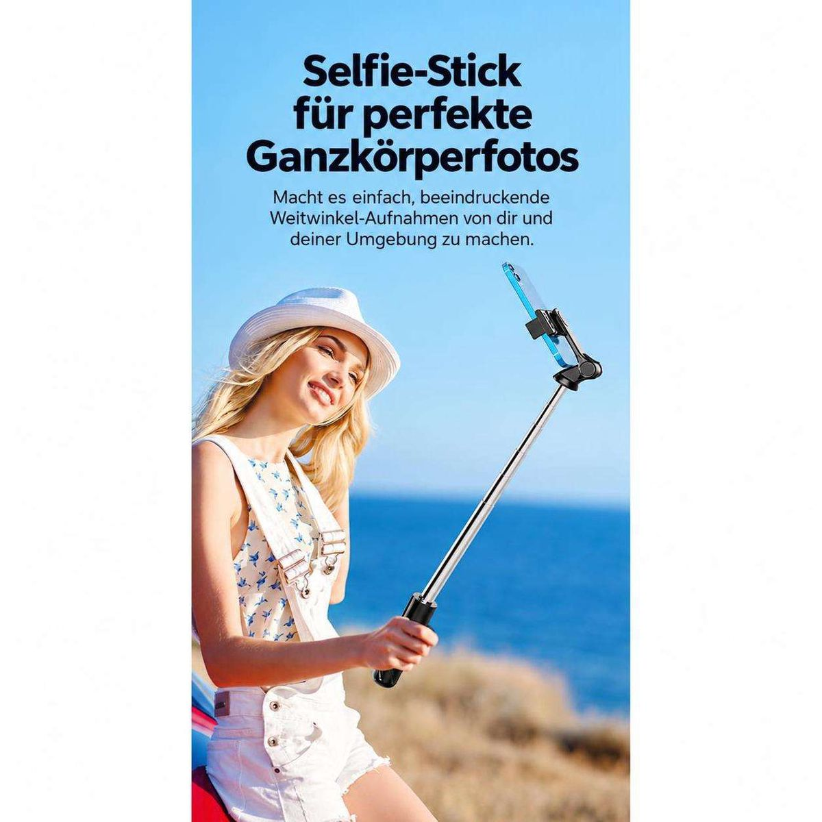 Eine blonde Frau in Hut und Overalls hält vor dem Meer einen langen Selfie-Stick mit einem Smartphone.