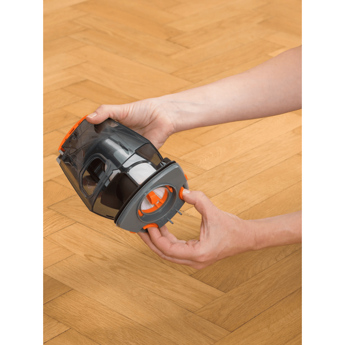 Manos sostienen un filtro de aspiradora gris y naranja sobre un piso de parquet.
