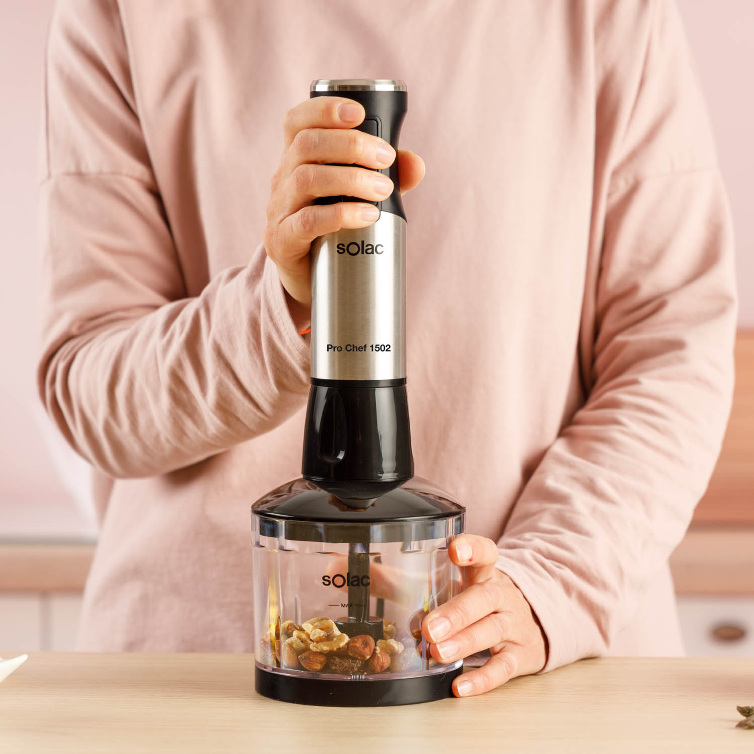 Osoba w różowej koszuli trzyma blender ręczny Solac nad miską z orzechami.