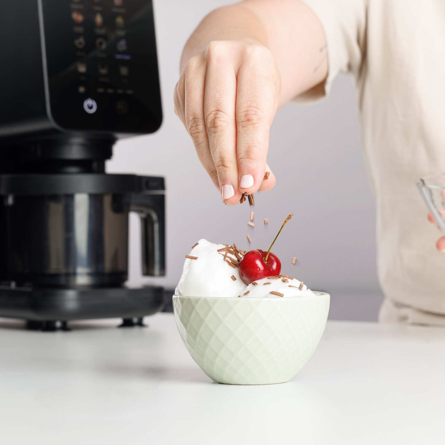 Osoba posypuje płatkami czekolady białe lody z wisienką, w tle ekspres do kawy.