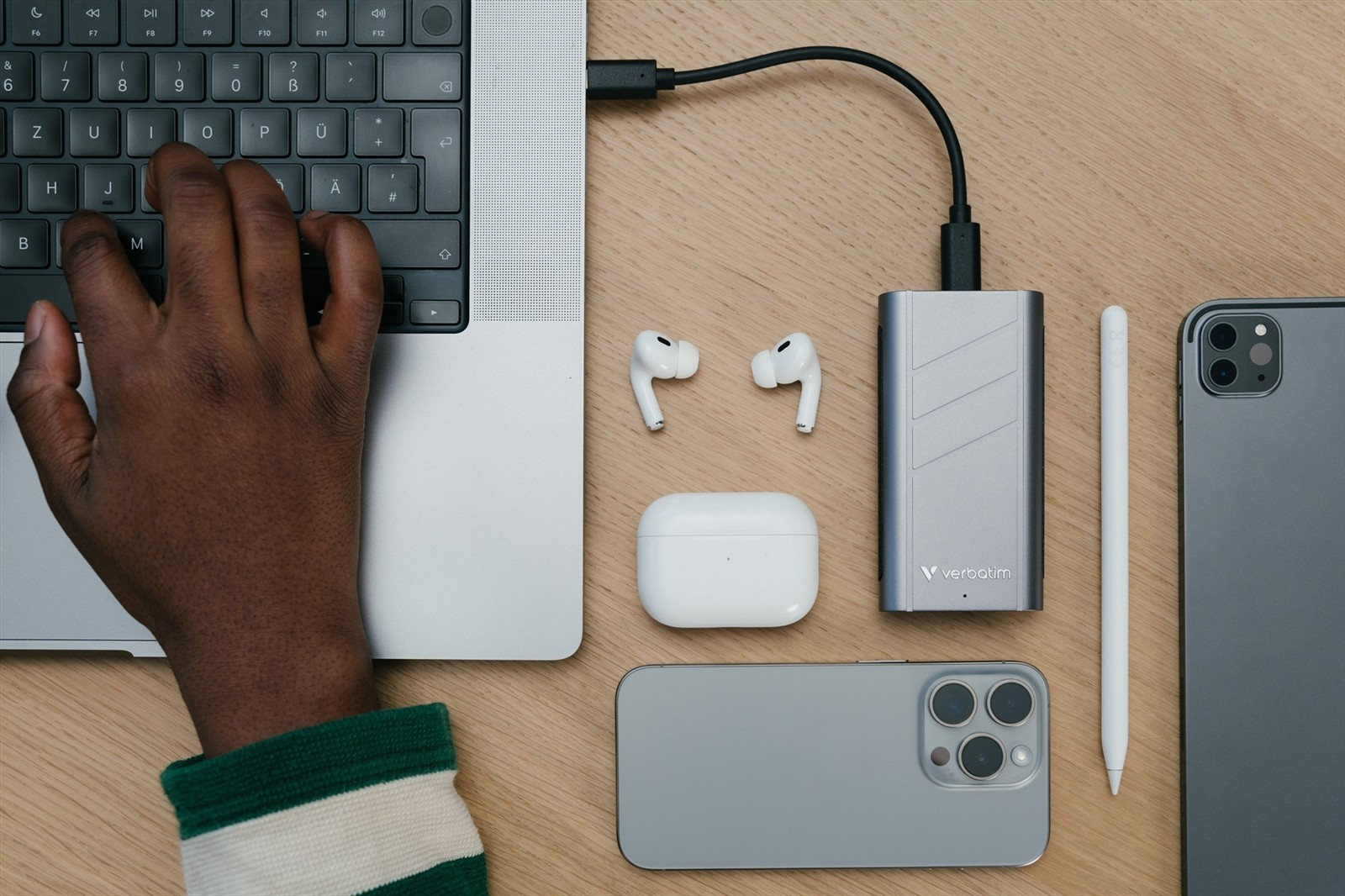 Mano escribiendo en portátil, con hub USB, AirPods, lápiz y dos móviles en madera.