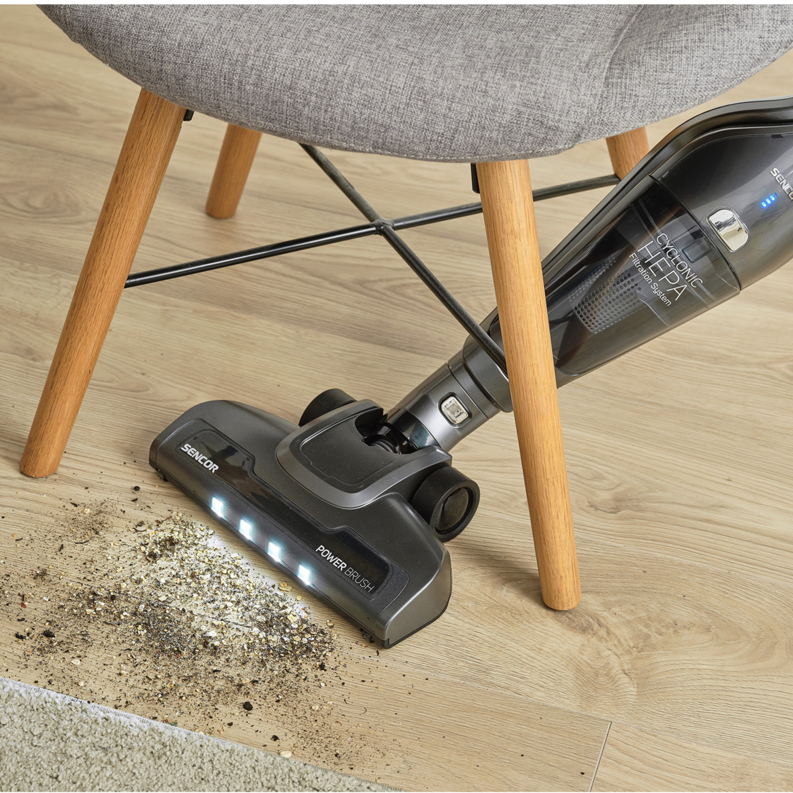 Una aspiradora inalámbrica gris limpia la suciedad en un piso de madera debajo de una silla.