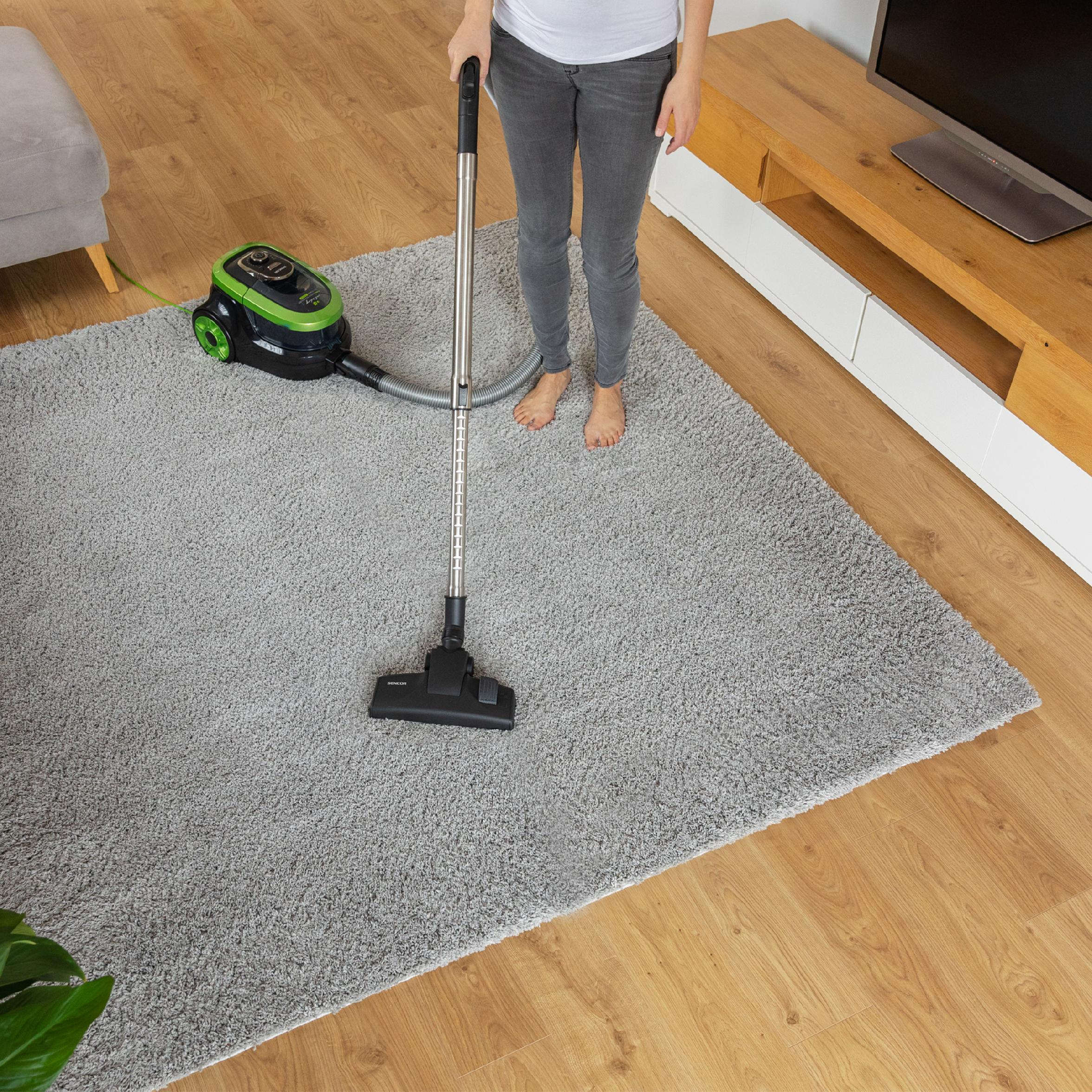 A woman vacuums a large grey carpet in a living room with wooden floors.