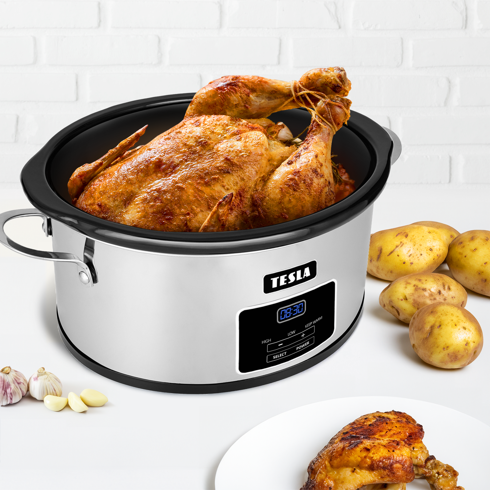 Un pollo intero arrosto in una pentola a cottura lenta Tesla, circondato da patate e aglio su sfondo bianco.
