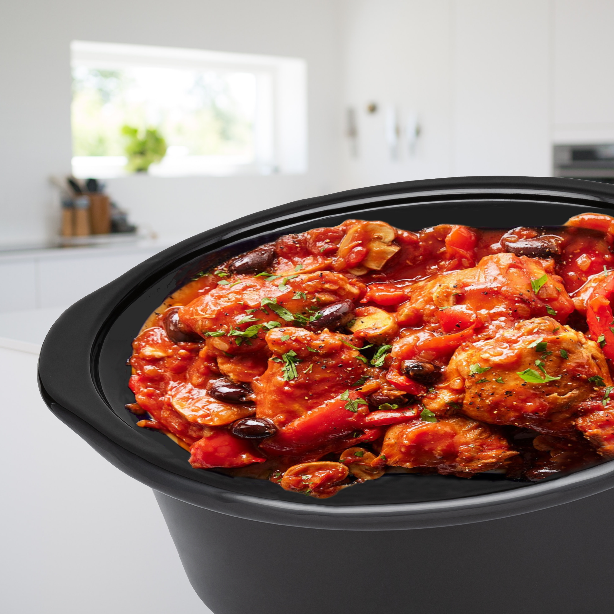Una pentola a cottura lenta nera piena di pollo al pomodoro con olive ed erbe, in una cucina luminosa.