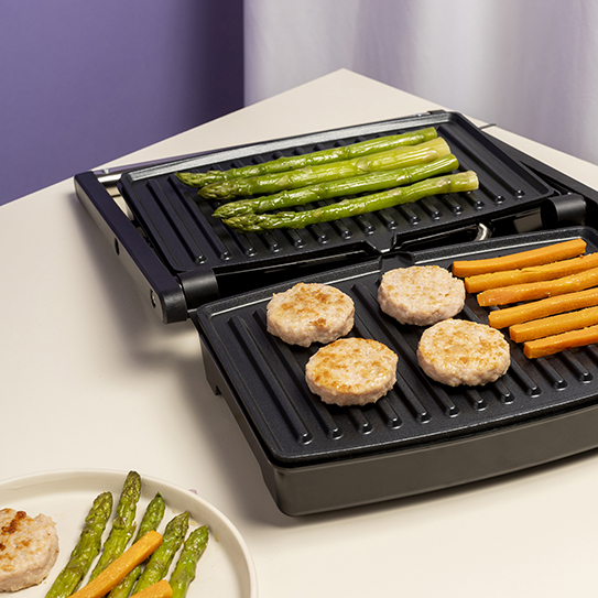 Otwarty grill kontaktowy ze szparagami, kotletami i paskami marchewki.