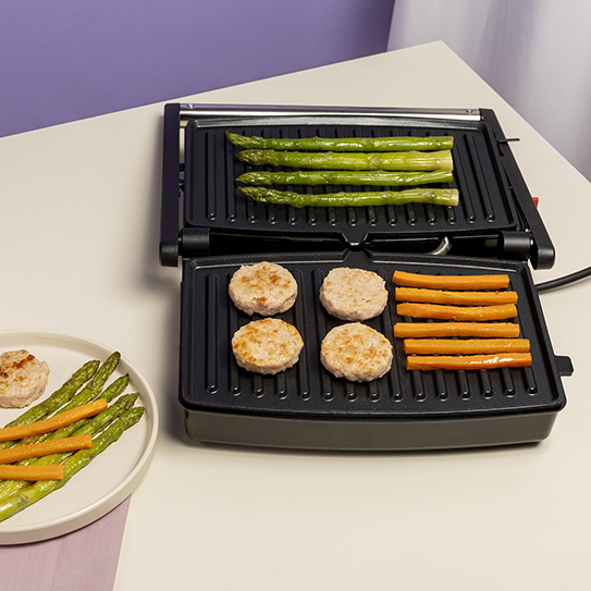 Otwarty grill stołowy z zielonym szparagami, okrągłymi kotletami i paskami marchewki oraz talerzem obok.