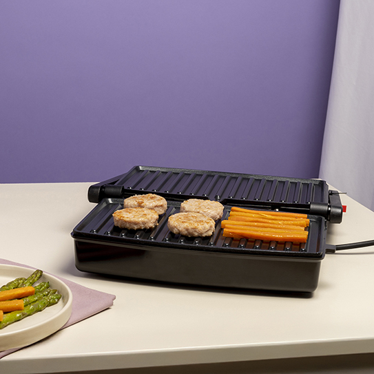 Grill elektryczny na stole gotuje kotlety do burgerów i słupki marchewki, z szparagami na talerzu.