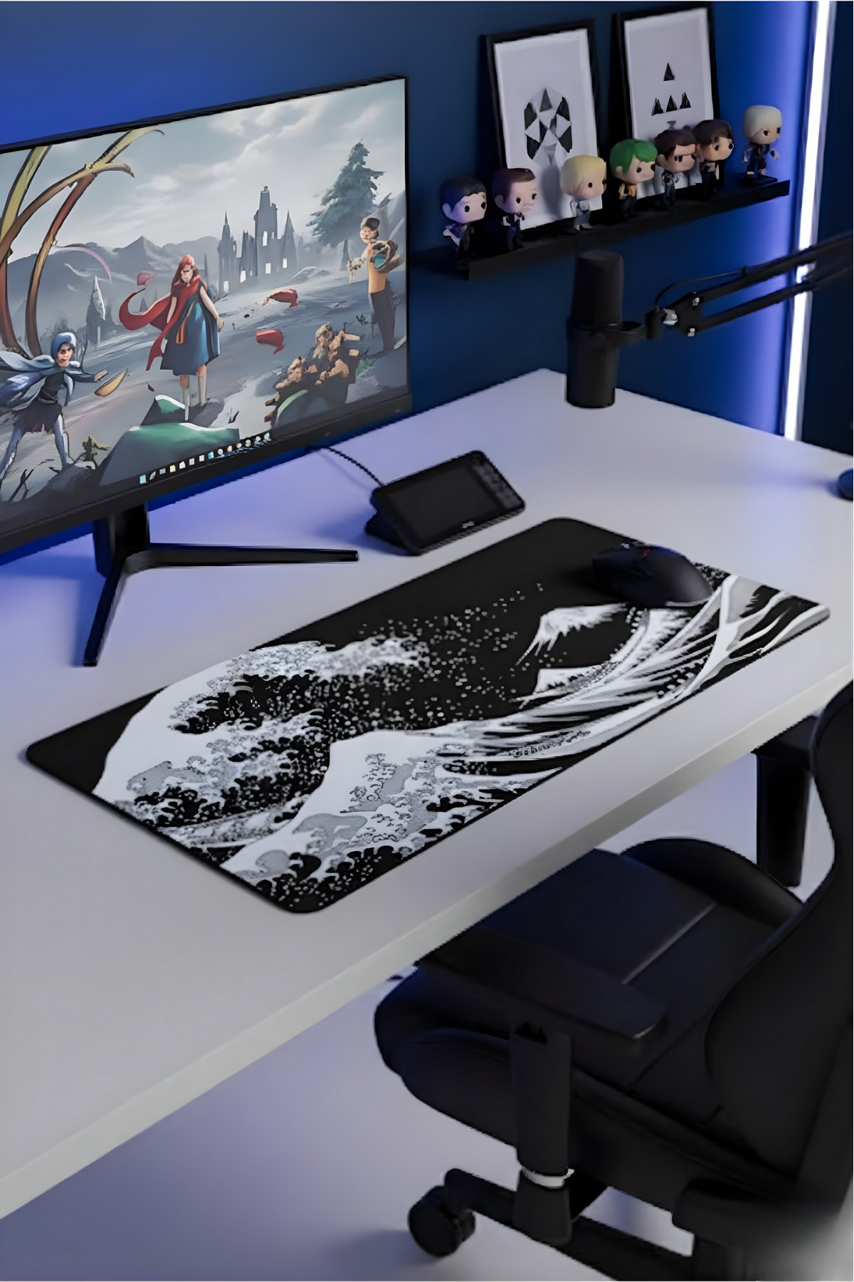 Mavi duvar önünde monitör, dalga mousepad, mikrofon ve figür rafı olan düzenli bir çalışma masası.