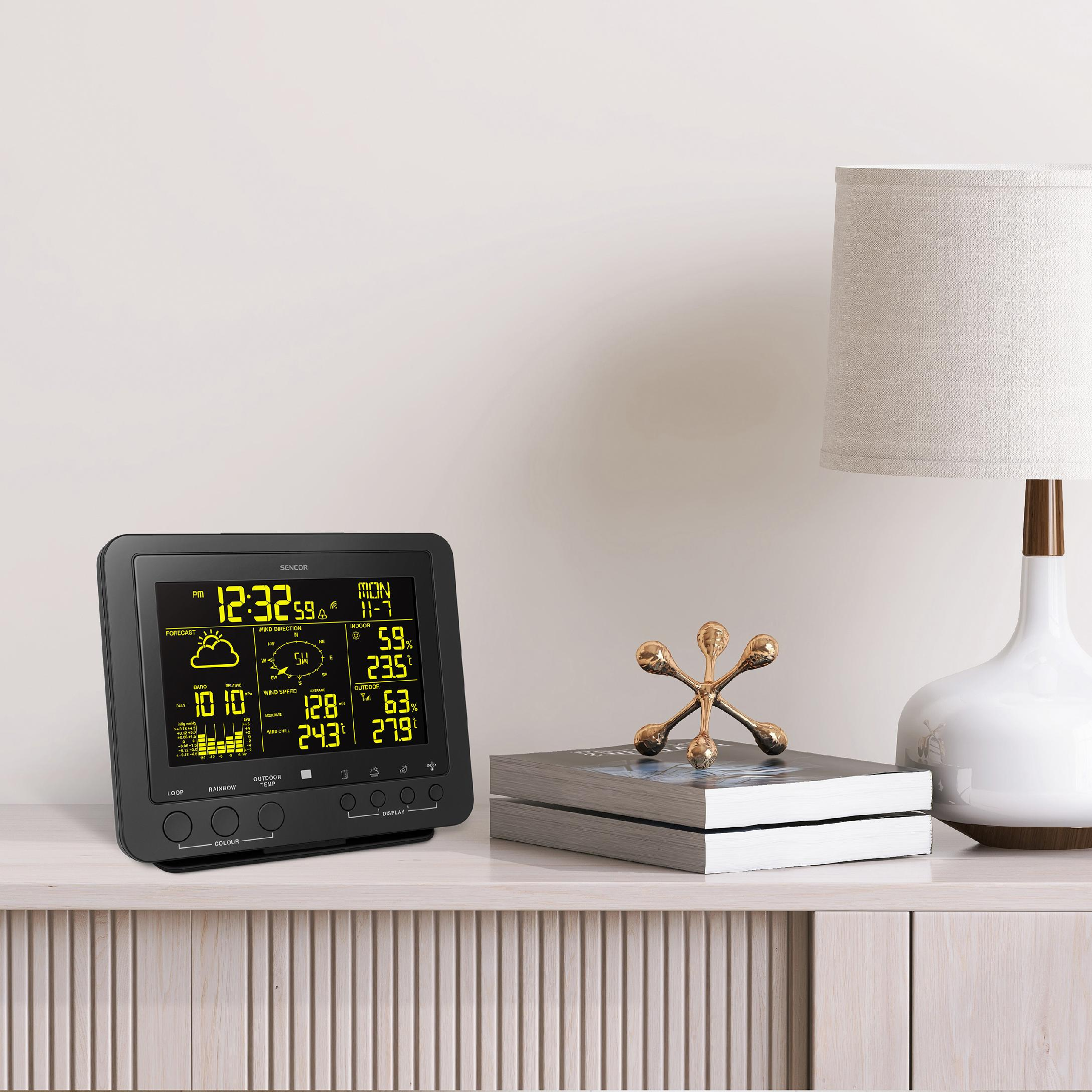 Eine schwarze Wetterstation mit gelbem Display steht auf einer Kommode neben gestapelten Büchern und einer Lampe.