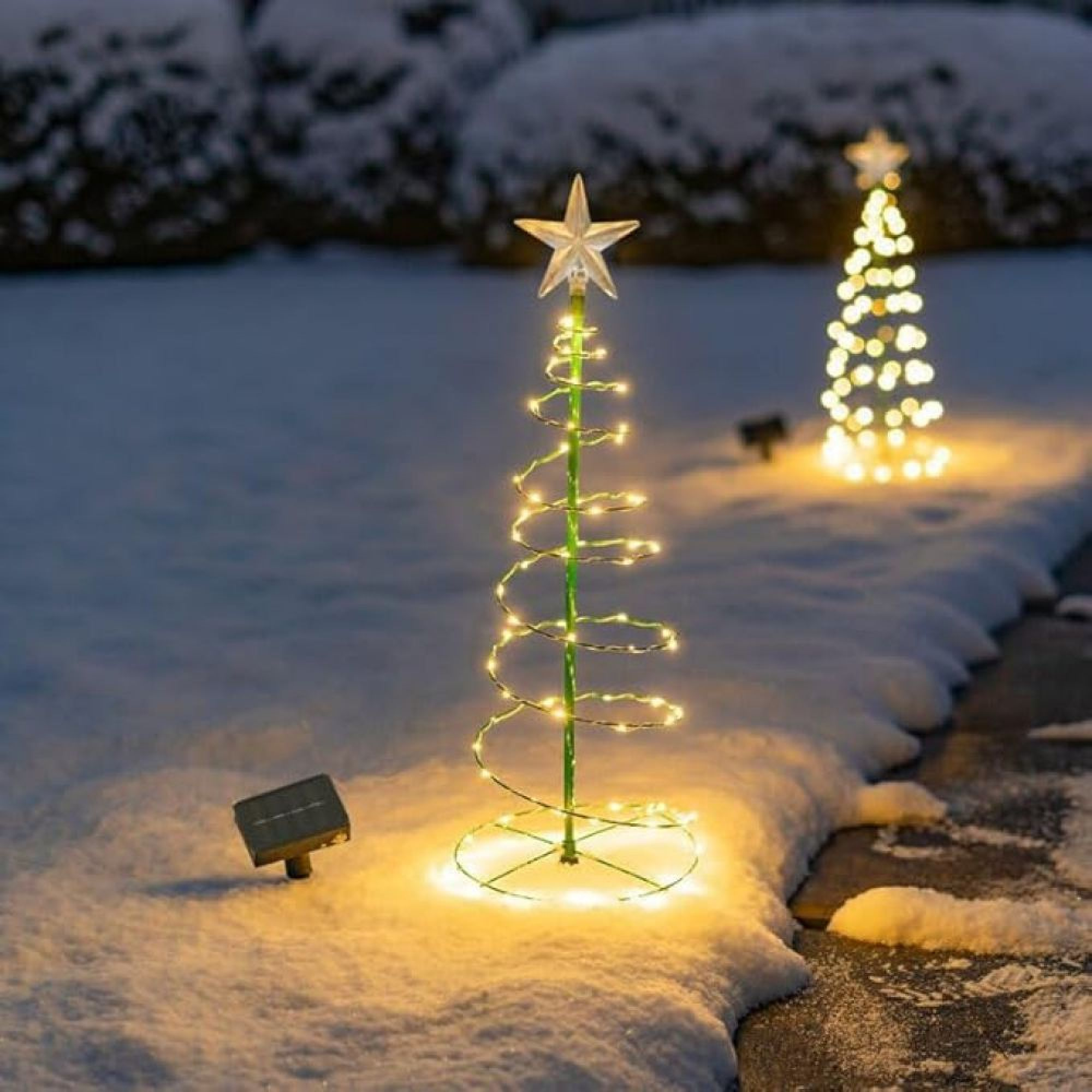 Zwei beleuchtete spiralförmige Weihnachtsbaum-Stecker stehen nachts in einem schneebedeckten Garten.