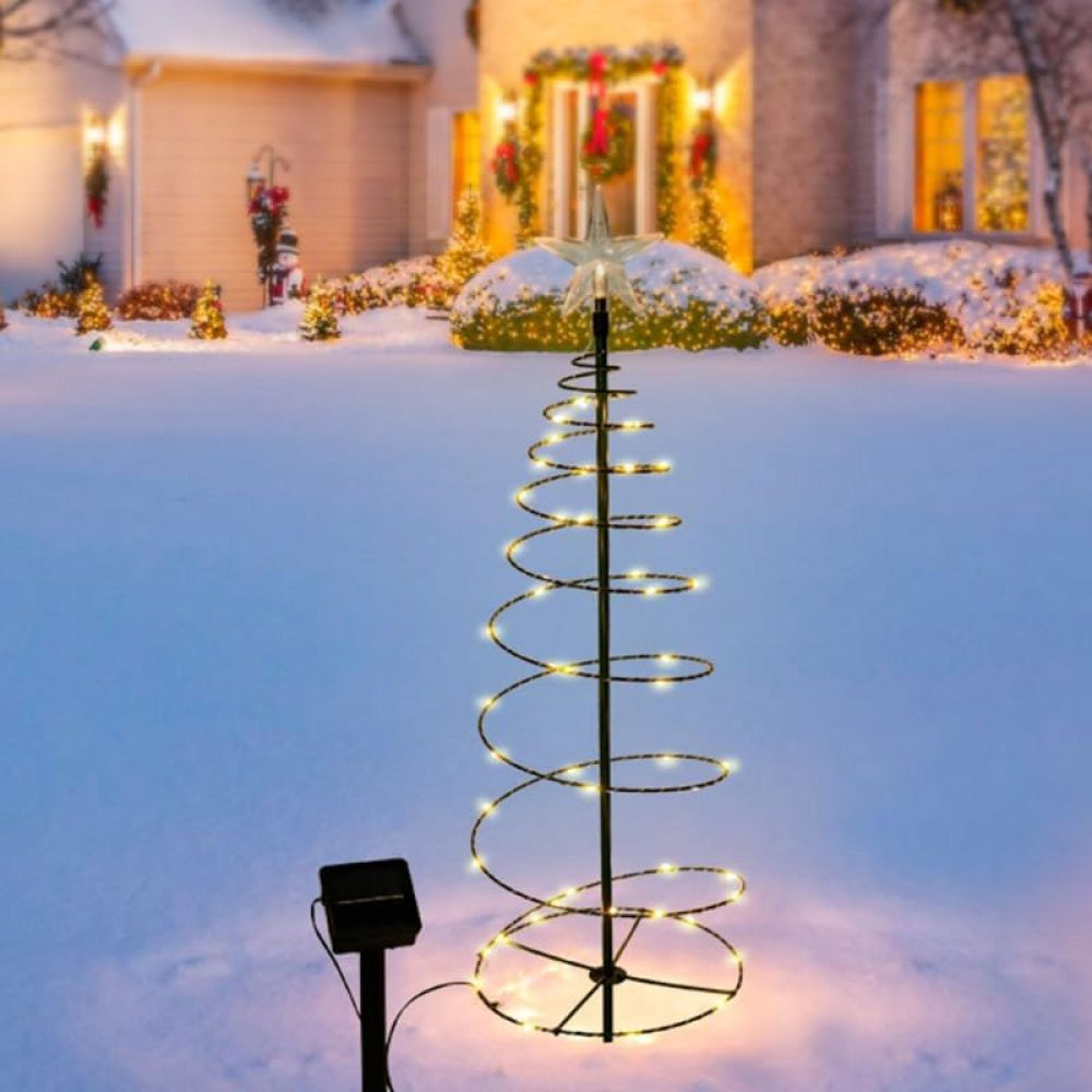 Beleuchteter spiralförmiger Weihnachtsbaum mit Stern im schneebezogenen Garten vor beleuchtetem Haus.
