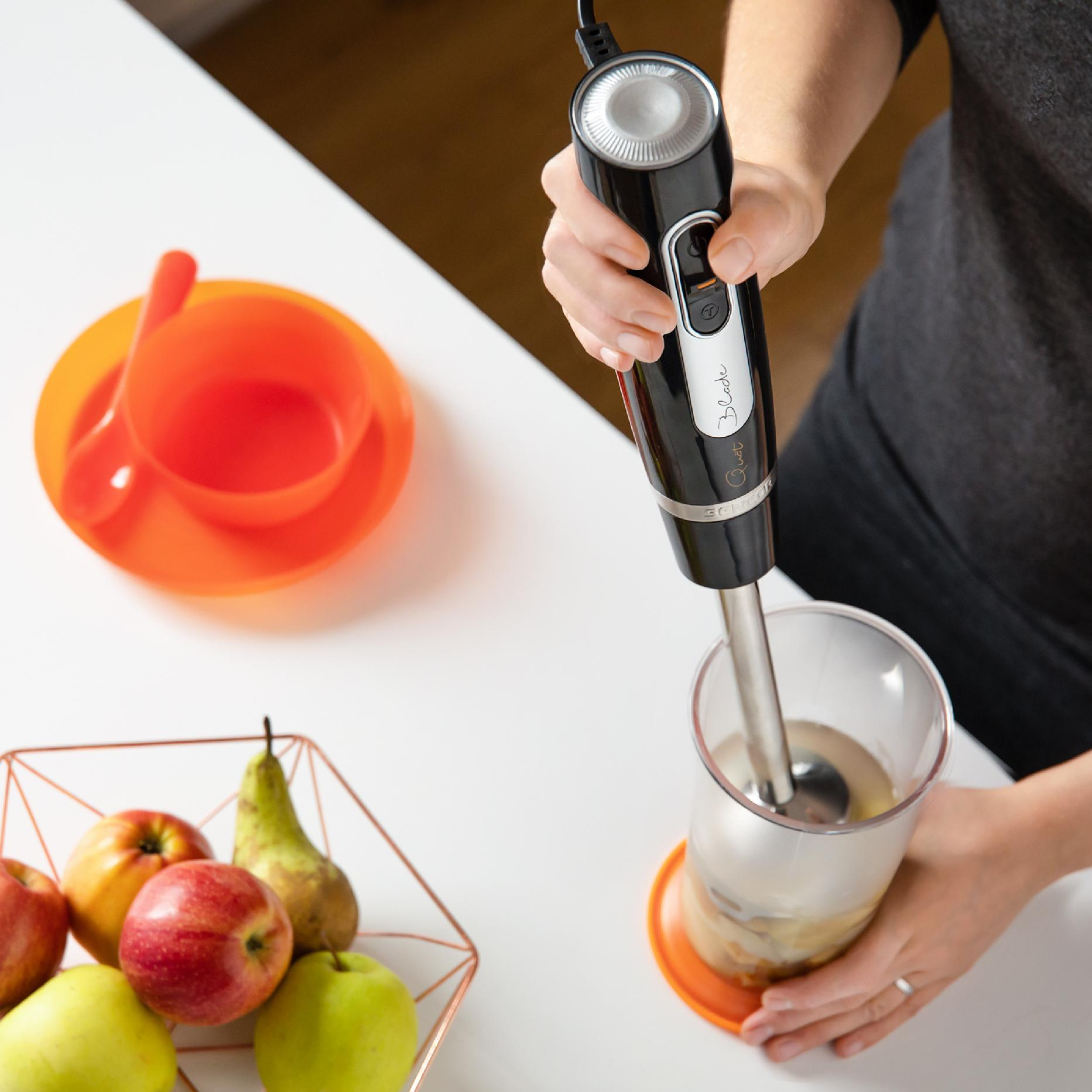 Eine Person nutzt einen schwarzen Stabmixer in einem Plastikbecher auf einem weißen Tisch, mit Äpfeln im Korb.