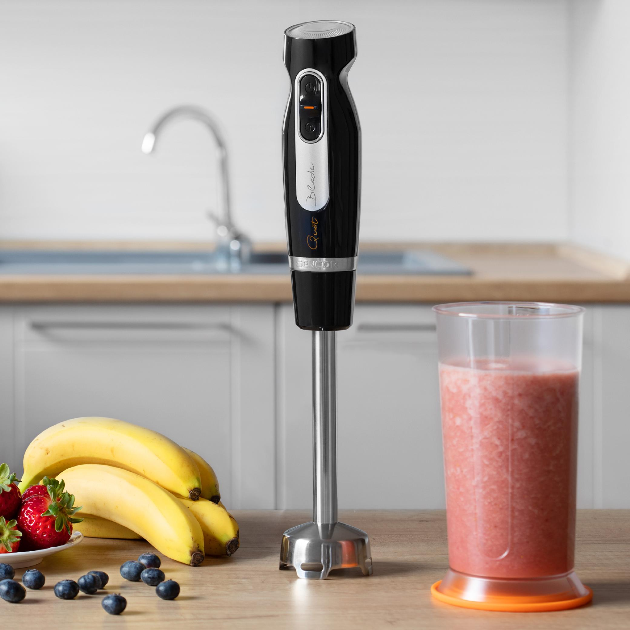 Ein schwarzer Stabmixer steht auf einem Holztisch mit Bananen, Erdbeeren, Blaubeeren und einem Smoothie.