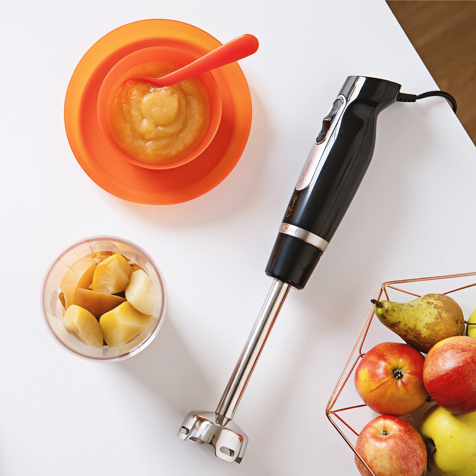 Ein schwarzer Stabmixer, eine Schüssel Apfelmark, Obststücke im Becher und ein Drahtkorb mit Äpfeln und Birnen auf weißem Tisch.