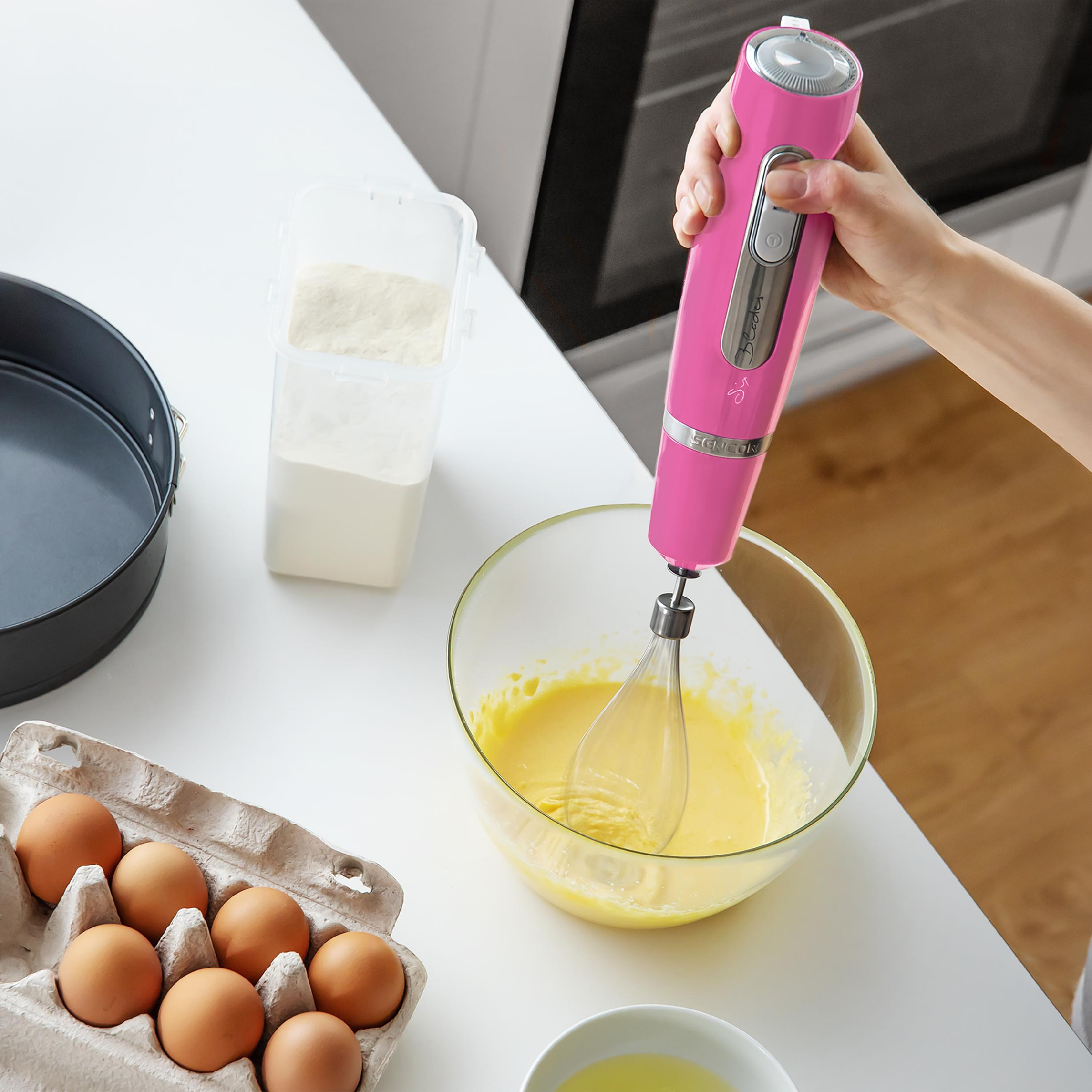Una mano sostiene una batidora rosa sobre masa en un bol de vidrio en una mesa blanca con huevos y harina.