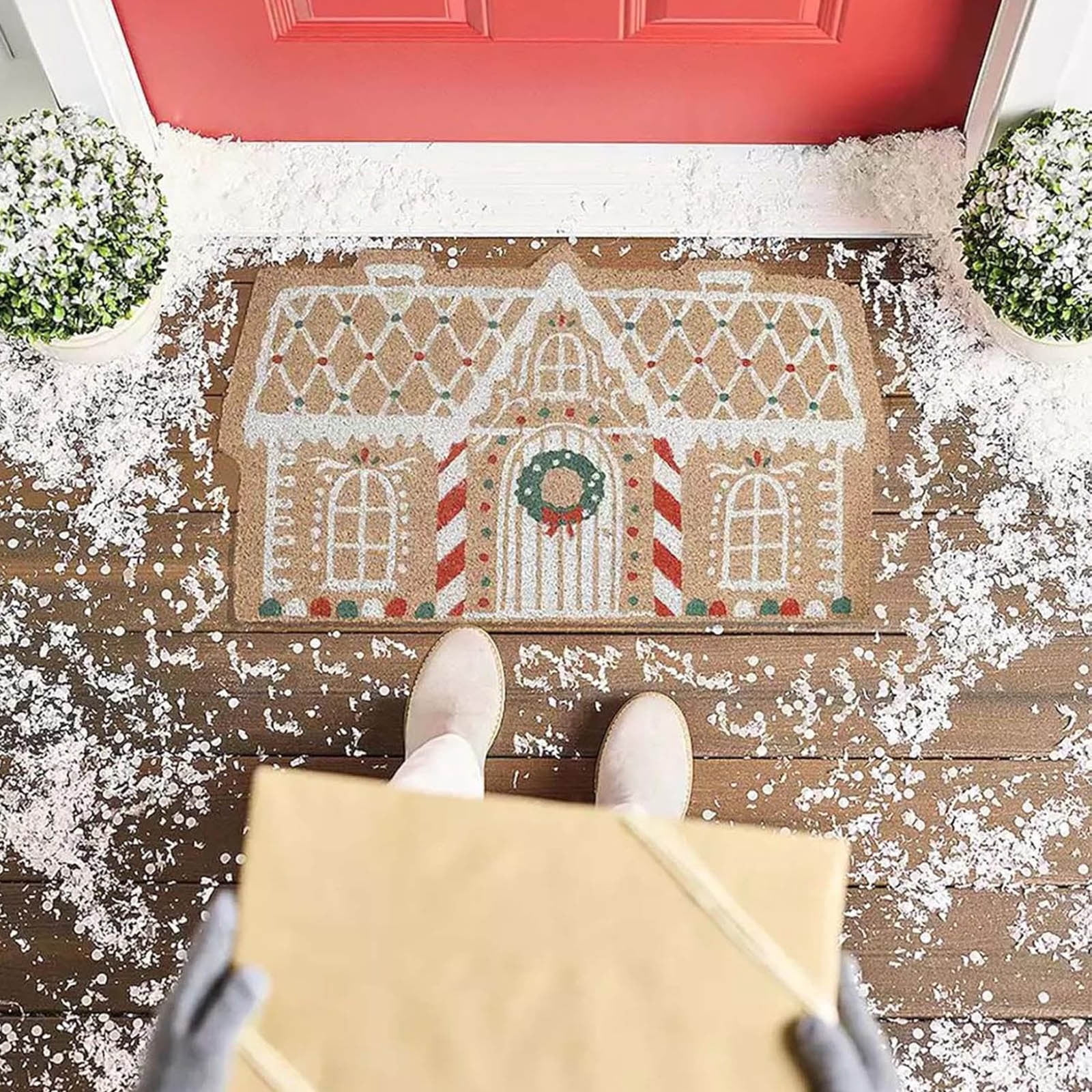 Person hält ein Paket vor einer Haustür auf einem fußabtreter mit Lebkuchenhausmotiv und Kunstschnee.