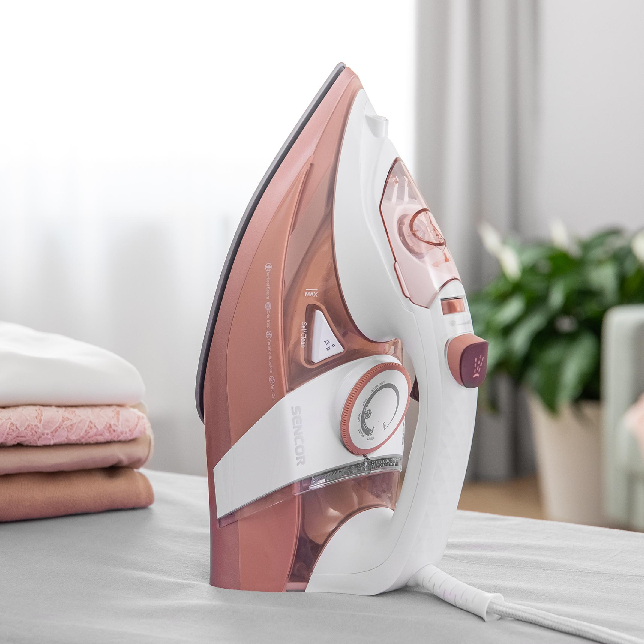 Una plancha de vapor blanca y oro rosa se encuentra sobre una superficie clara, al lado de ropa doblada.